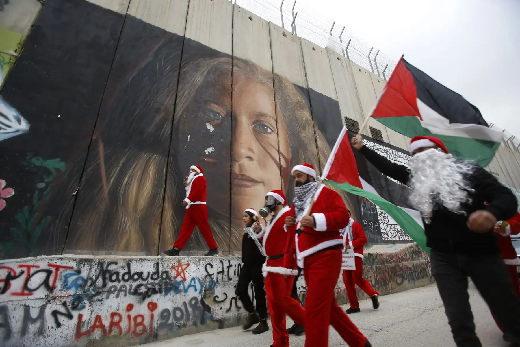 Palestinos vestidos como Papá Noel en Belén, cuelgan carteles en una pintura mural del militante palestino Ahed Tamimi en la polémica barrera de separación de Israel en la ciudad Cisjordana de Belén. Foto: AFP