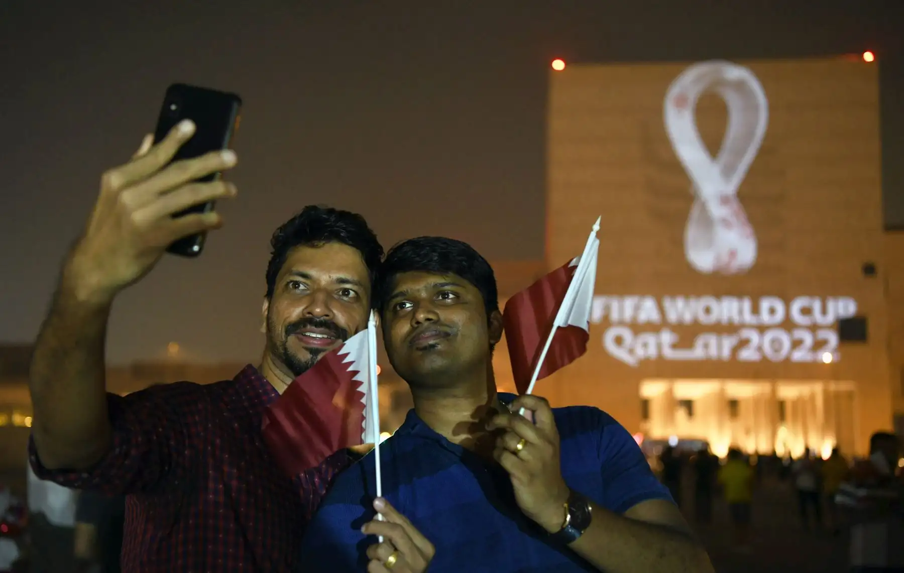 La gente hace selfies en el mercado tradicional Souq Waqif de Doha, mientras que el logotipo oficial de la Copa Mundial de la FIFA Qatar 2022 se proyecta en el frente de un edificio.Foto:AFFP