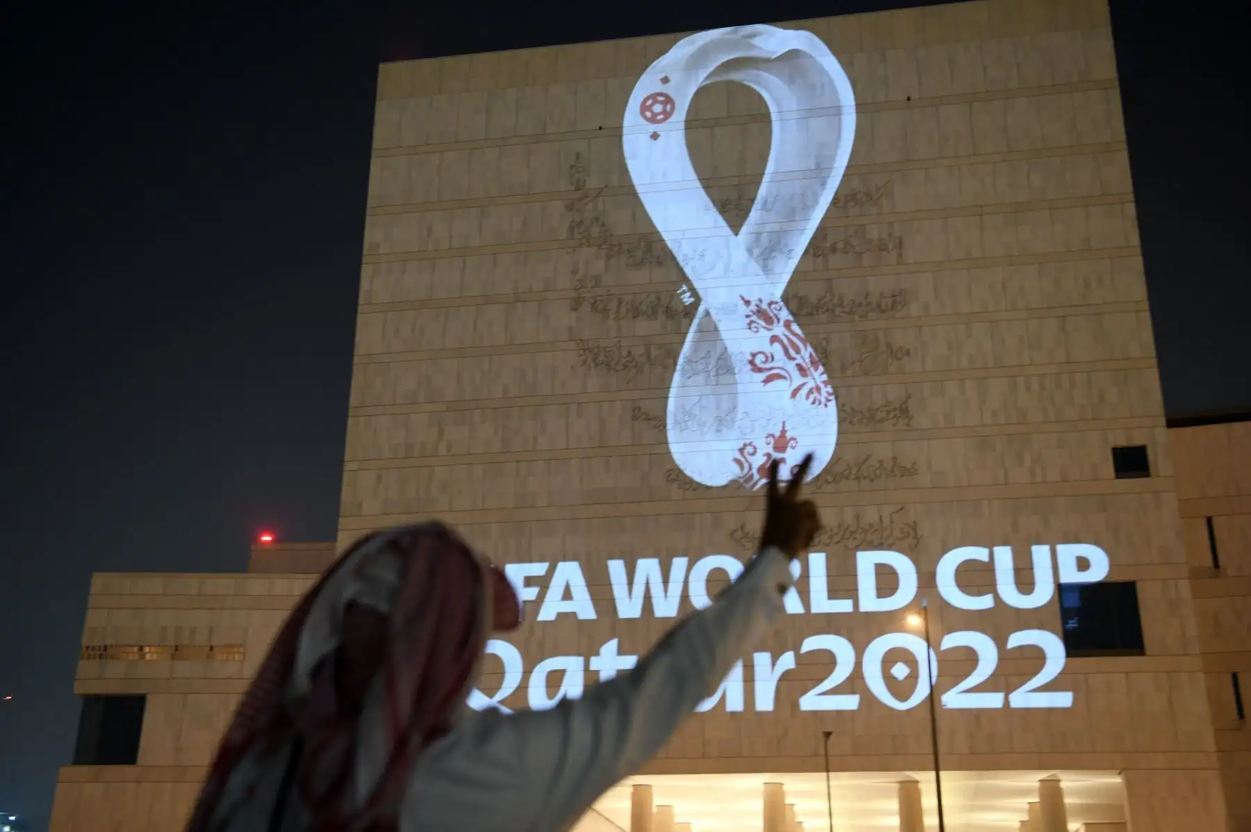 Un hombre qatarí hace un gesto mientras la gente se reúne en el mercado tradicional Souq Waqif de Doha, la capital, mientras que el logotipo oficial de la Copa Mundial de la FIFA Qatar 2022 se proyecta en la parte delantera de un edificio .Foto:AFP