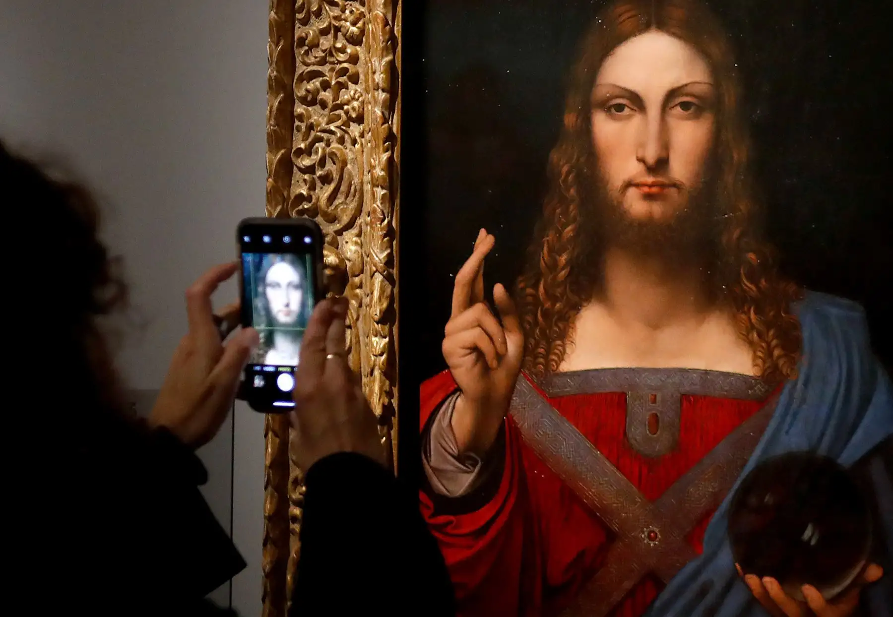 Una persona toma una fotografía con un teléfono móvil en una pintura al óleo de "Salvator Mundi" de Leonardo da Vinci, durante la inauguración de la exposición "Leonardo da Vinci", el 22 de octubre de 2019 en el museo del Louvre. Foto: AFP