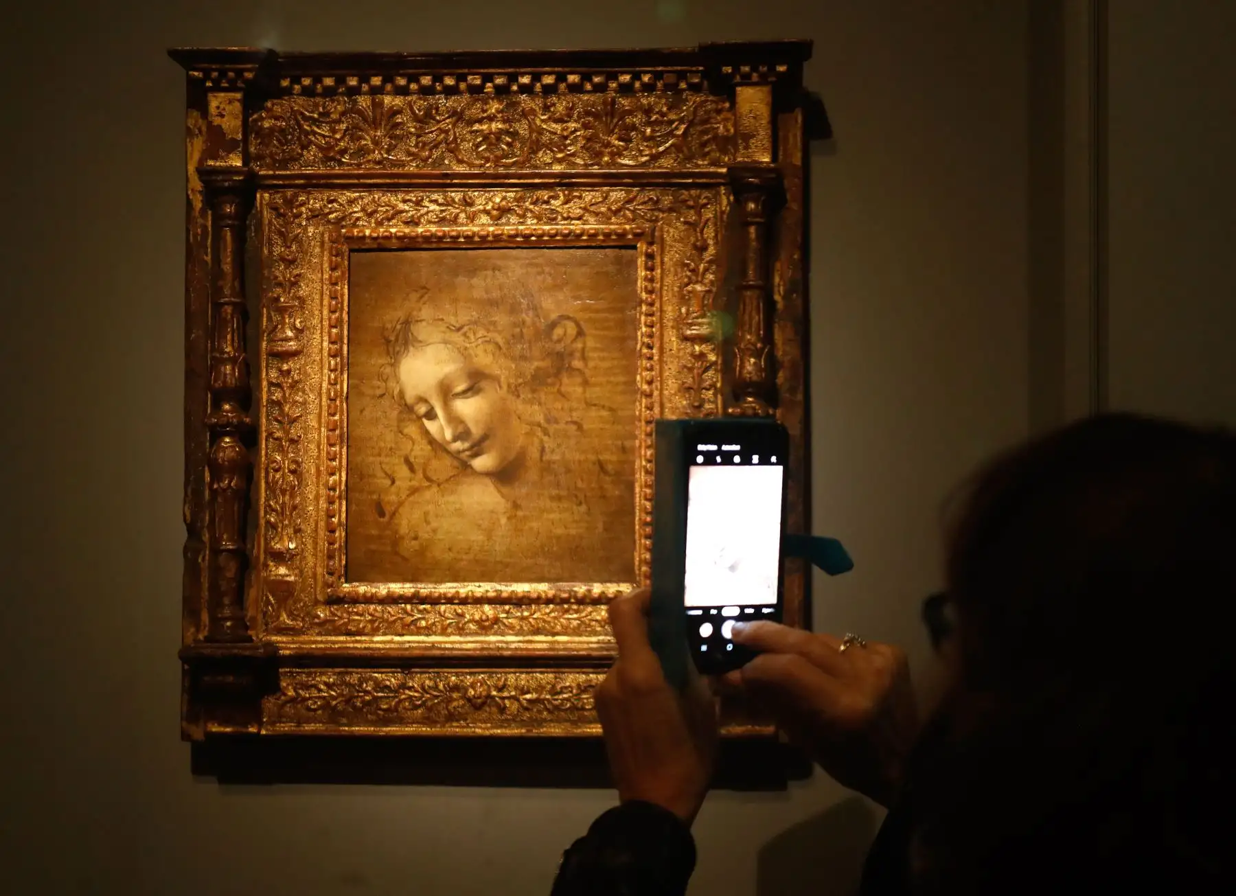 Una persona toma una fotografía con un teléfono móvil en una pintura al óleo sobre madera de "La cabeza de una mujer" de Leonardo da Vinci, también conocida como La Scapigliata, durante la inauguración de la exposición "Leonardo da Vinci". Foto: AFP