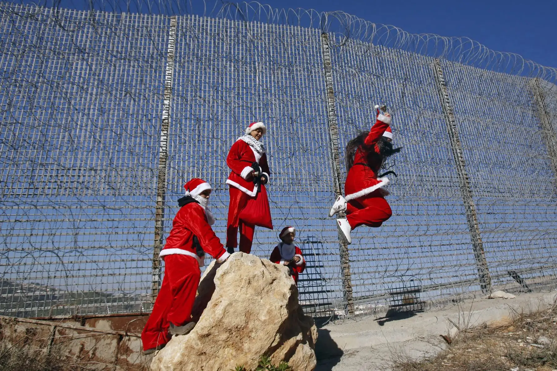 Los palestinos vestidos con trajes de Navidad saltan de una roca por una sección de alambre de púas de la controvertida barrera de separación de Israel cerca de la aldea de Wallajeh, al oeste de la ciudad bíblica de Belén en la Cisjordania ocupada.
Foto: AFP