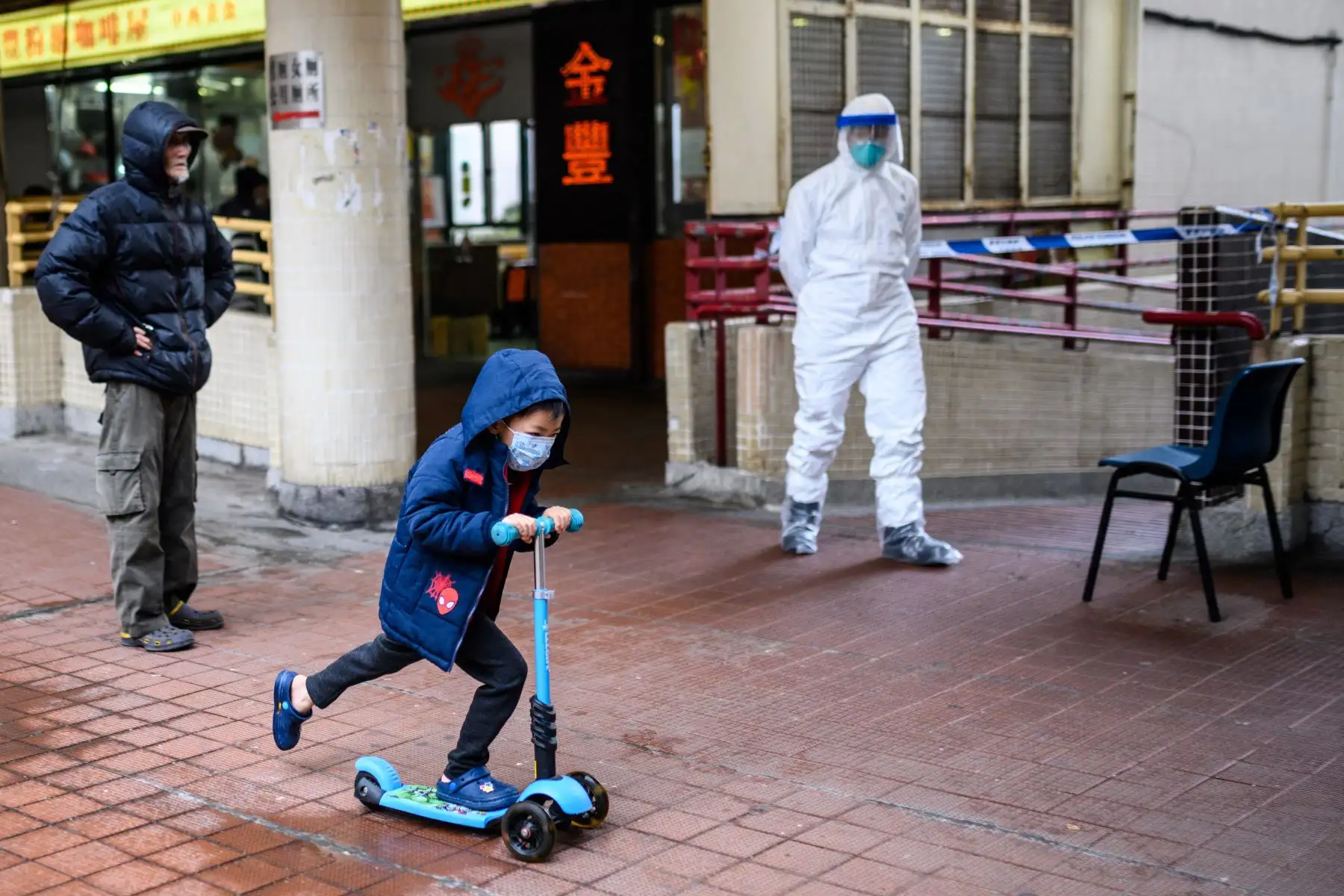 Un niño con máscara facial pasea en scooter frente a un oficial de policía que usa equipo de protección afuera de la casa Hong Mei en  Hong Kong. AFP