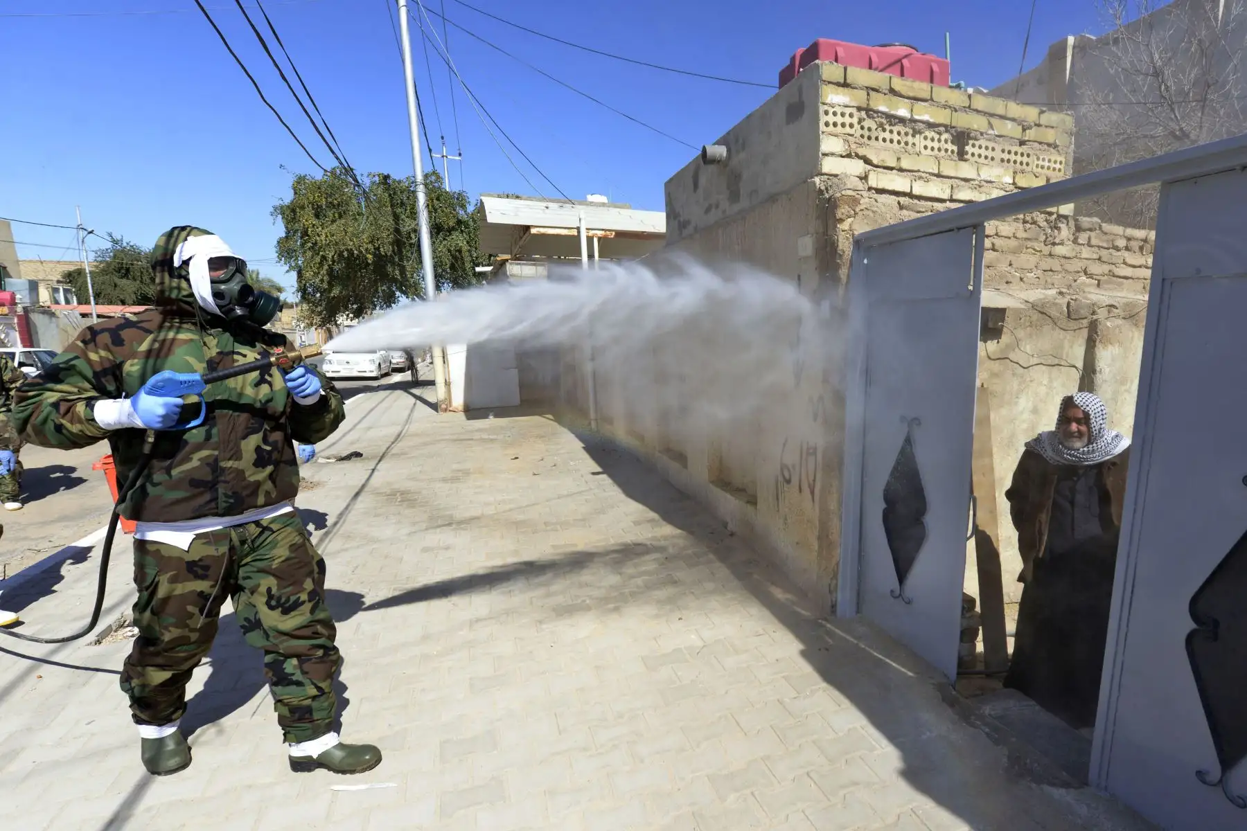 Un hombre iraquí mira por una puerta de entrada mientras un miembro de la defensa civil desinfecta un vecindario donde se confirmó un caso reciente de infección por coronavirus. Foto: AFP