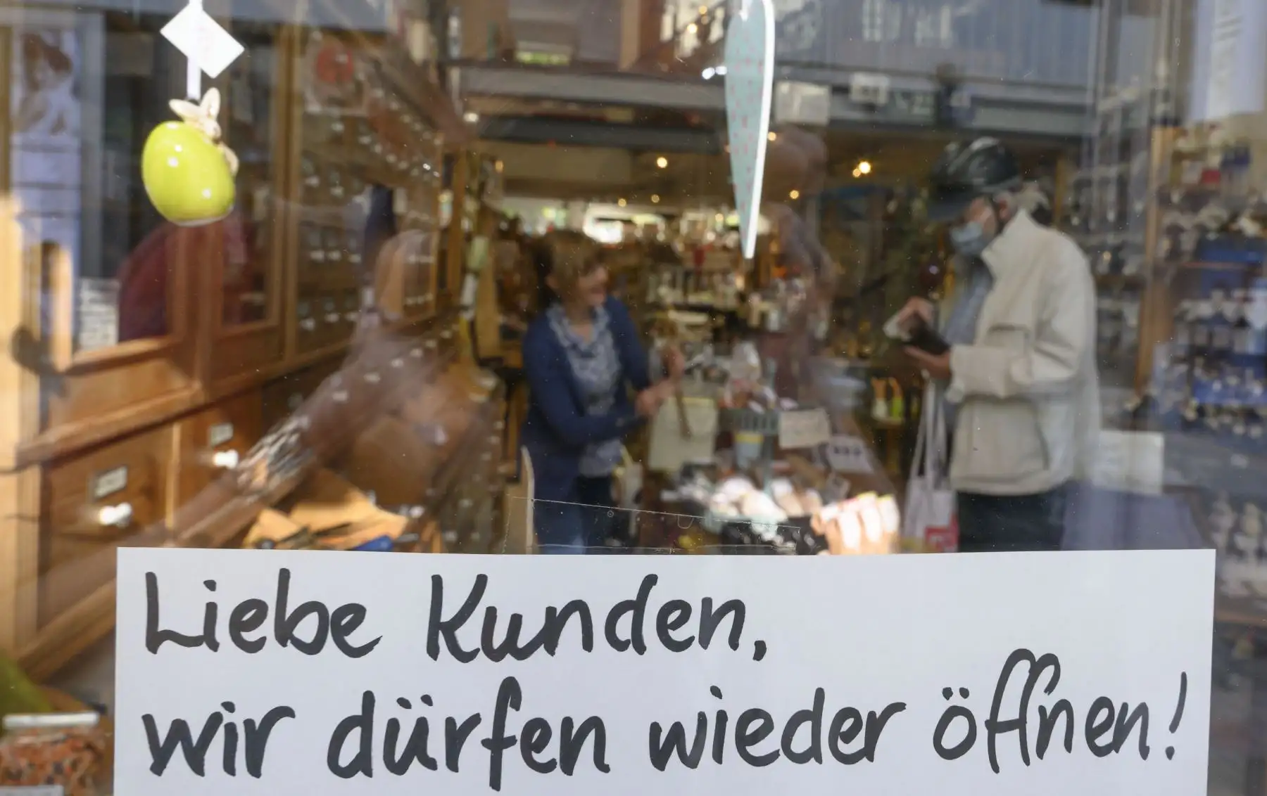 Elisabeth Merzach, dueña de una tienda de té y vino, se para detrás de un mostrador mientras habla con un cliente; además se muestra un cartel en el escaparate que dice "Estimados clientes, ¡podemos abrir de nuevo!" en Ludwigsburg, sur de Alemania, el 20 de abril de 2020, en medio de la nueva pandemia de coronavirus COVID-19. Foto: AFP