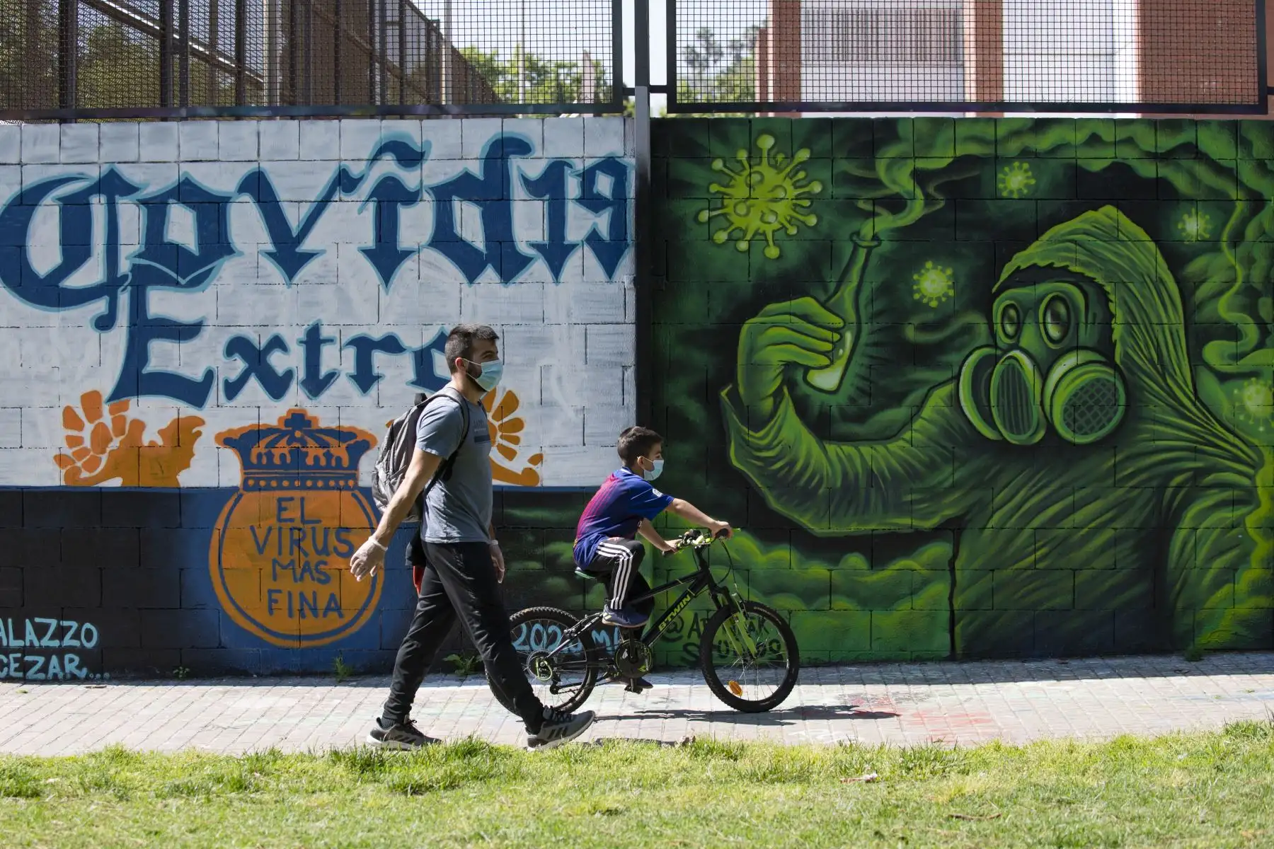 Un niño monta su bicicleta junto a su padre mientras pasan frente a un graffiti COVID-19 en Barcelona el 26 de abril de 2020. Foto: AFP