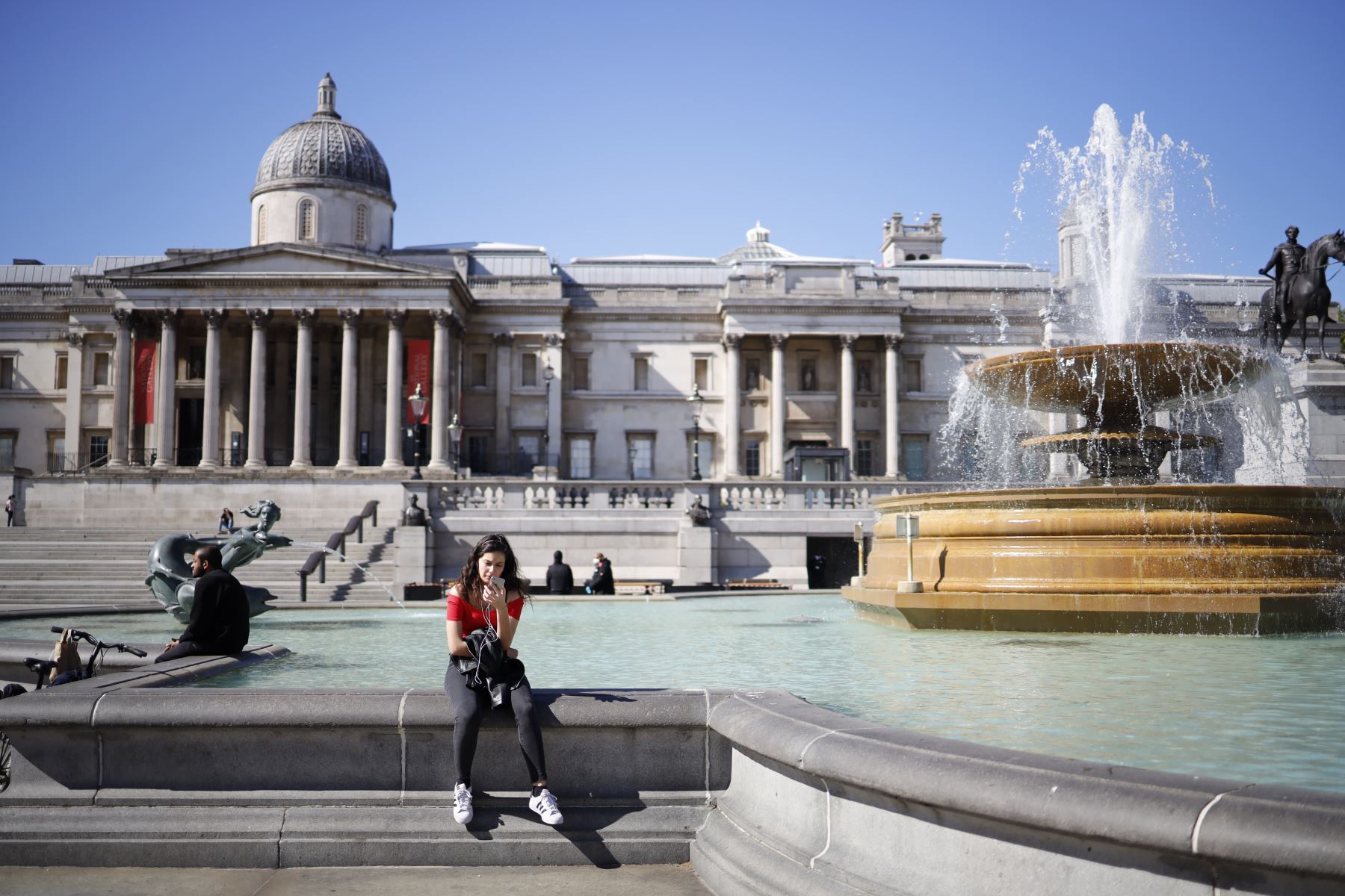 Una mujer se sienta en el borde de la fuente en Trafalgar Square en la Galería Nacional en el fondo en Londres. Foto: AFP