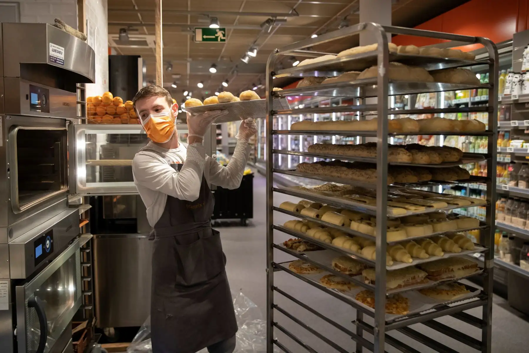Un empleado que usa guantes y una máscara protectora de boca y nariz coloca bandejas con masa en un horno en un supermercado Franprix. Foto: AFP