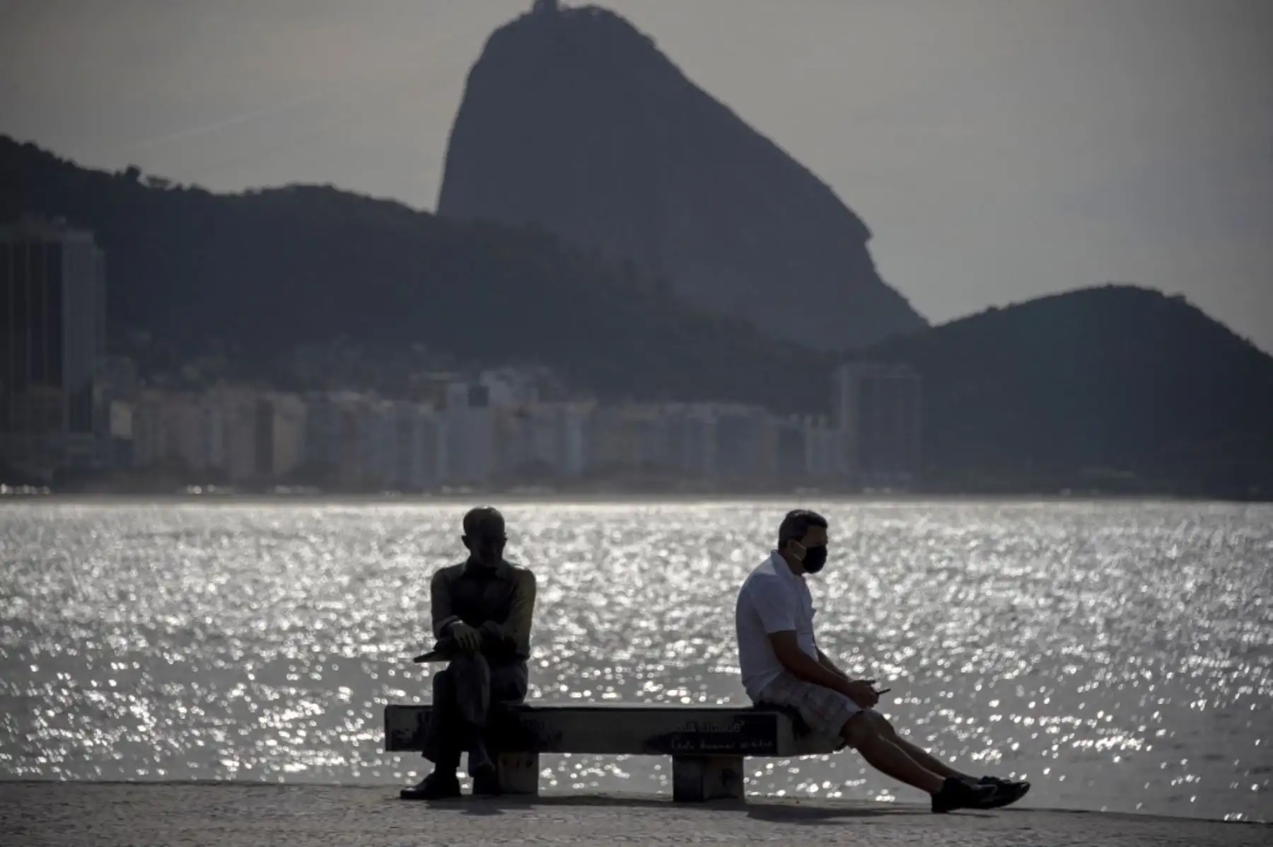 Un hombre con una máscara facial se sienta junto a la estatua del escritor brasileño Carlos Drummond de Andrade (L) en el paseo marítimo de la playa de Copacabana en el estado de Río de Janeiro. Foto: AFP