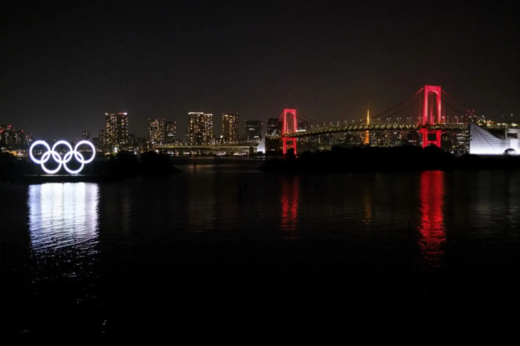 Los anillos olímpicos se ven junto con el Puente del Arco Iris iluminado en rojo por la noche, después de que el gobierno de Tokio decidió emitir una alerta debido a un aumento en los casos de coronavirus COVID-19 en Tokio. Foto: AFP