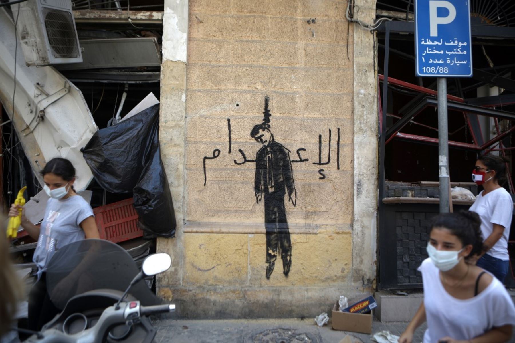 Miembros de la sociedad civil libanesa que llevan escobas usadas para limpiar escombros, pasan junto a una pintura mural que representa a un político ahorcado en el vecindario de Mar Mikhael, Beirut, parcialmente dañado. Foto: AFP