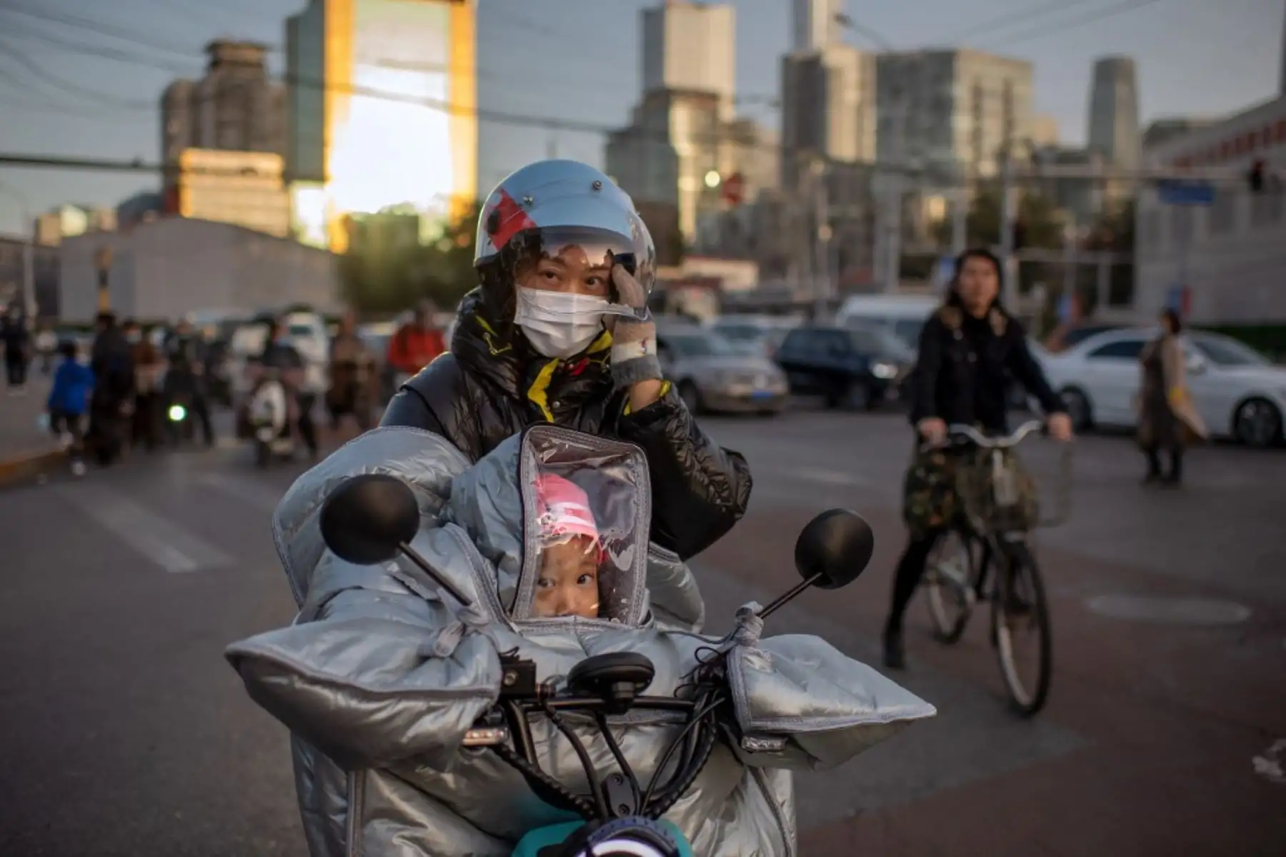 Una mujer que usa una mascarilla como medida preventiva contra el coronavirus covid-19 espera para cruzar una calle con su hija mientras ambas montan en una bicicleta eléctrica durante la hora pico en Beijing. Foto: AFP
