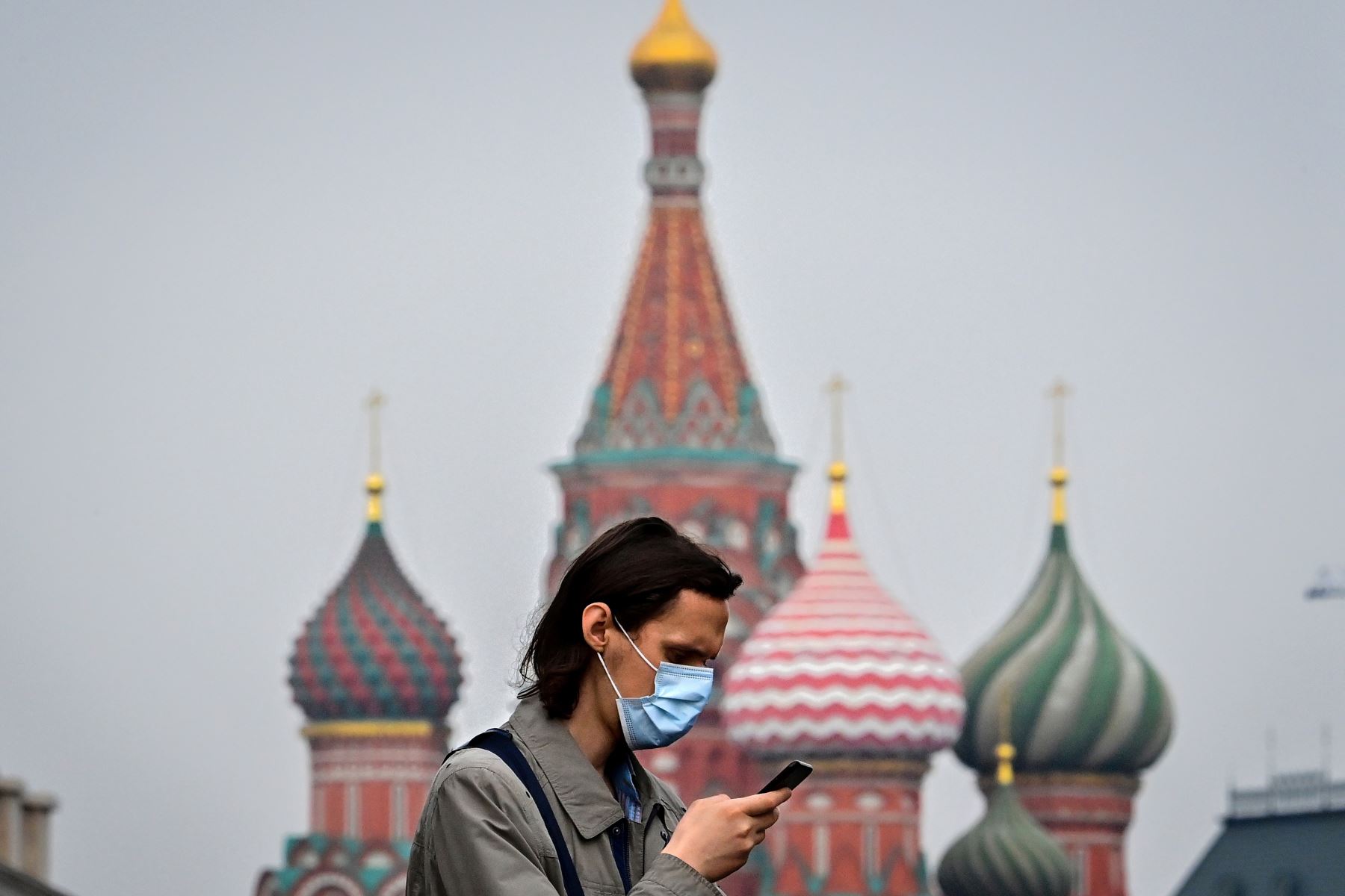Un hombre que usa una mascarilla para protegerse contra la enfermedad del coronavirus usa su teléfono móvil mientras transita frente a la Catedral de San Basilio, en el centro de Moscú. Foto: AFP