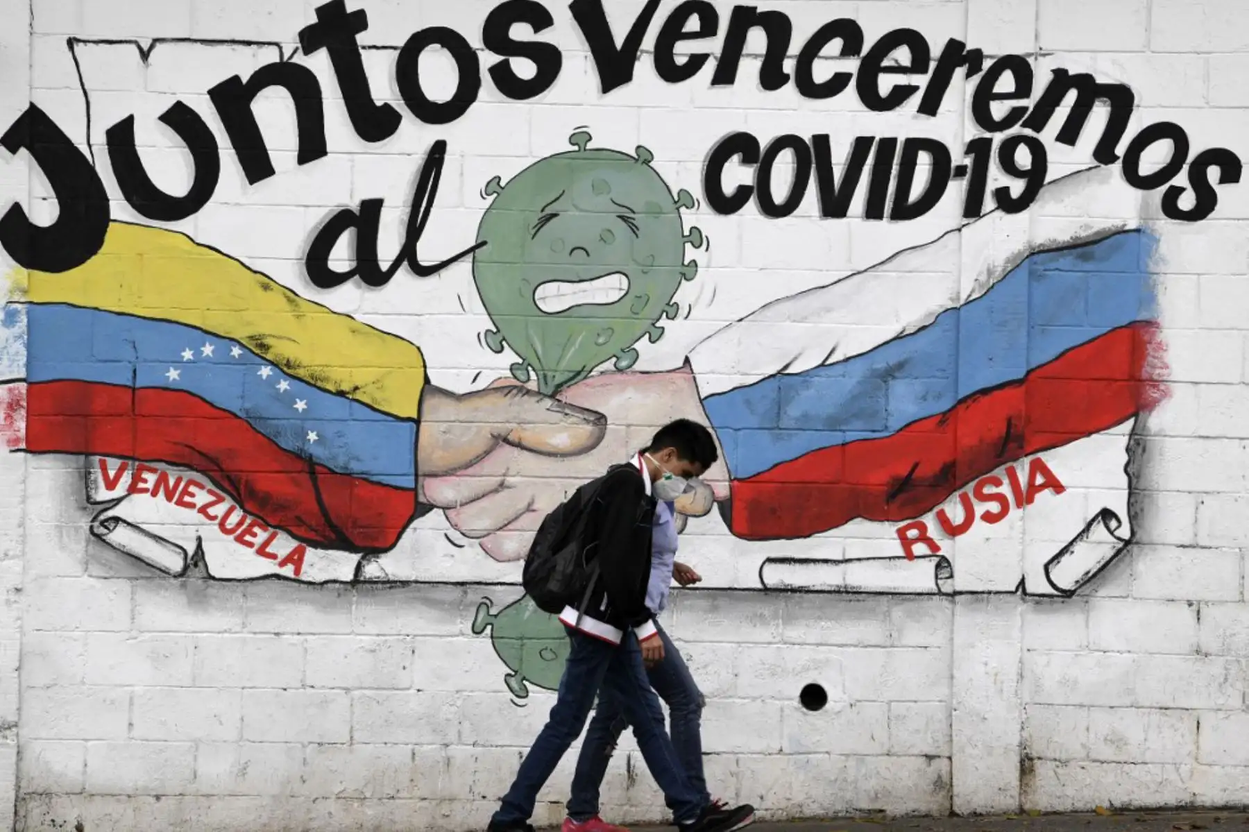 Jóvenes pasan frente a un grafiti con las banderas de Venezuela y Rusia que dice "Juntos derrotaremos al COVID-19" en Caracas.

Foto: AFP