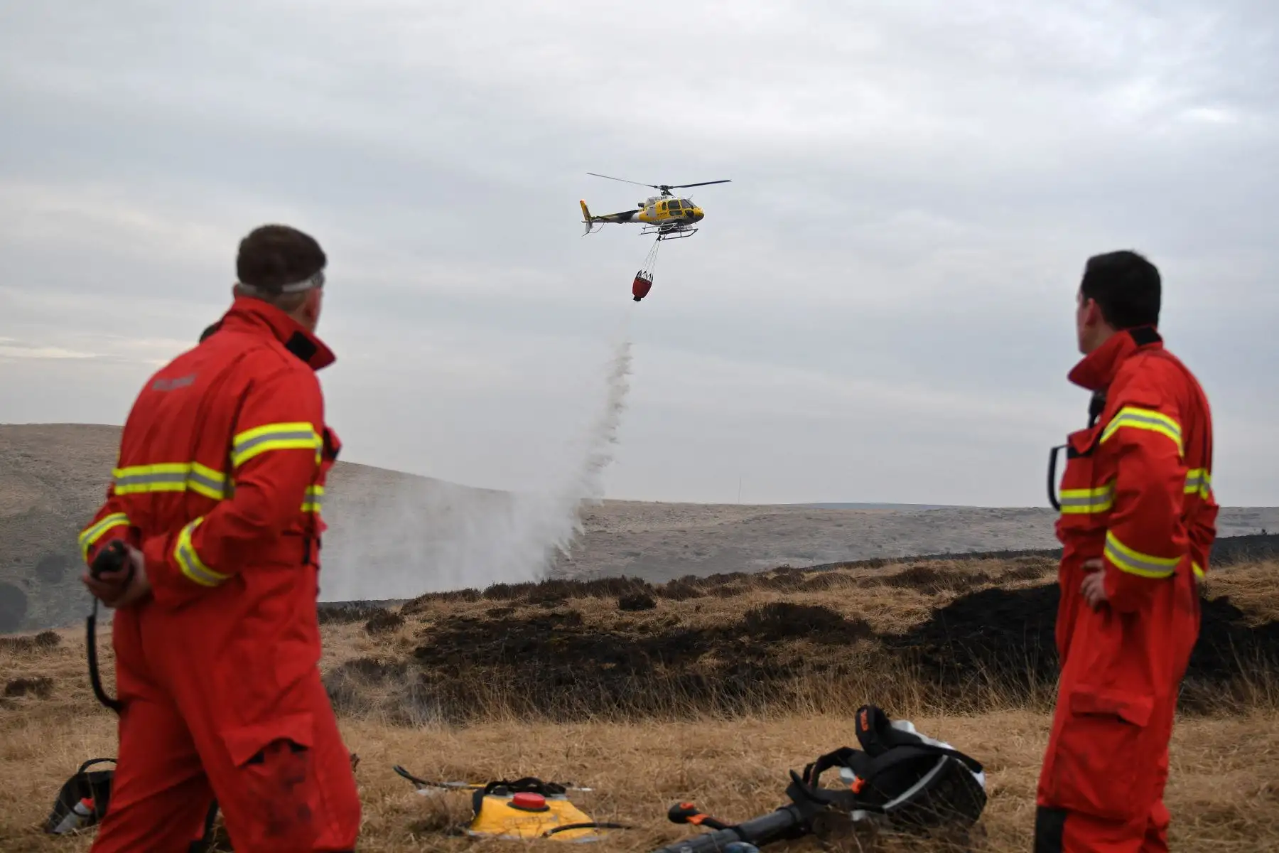 Los bomberos observan cómo un helicóptero arroja agua, tomada de un embalse cercano, después que resurgiera el incendio del páramo en Marsden Moor, cerca de Huddersfield, en el norte de Inglaterra, el 26 de abril de 2021. Foto: AFP