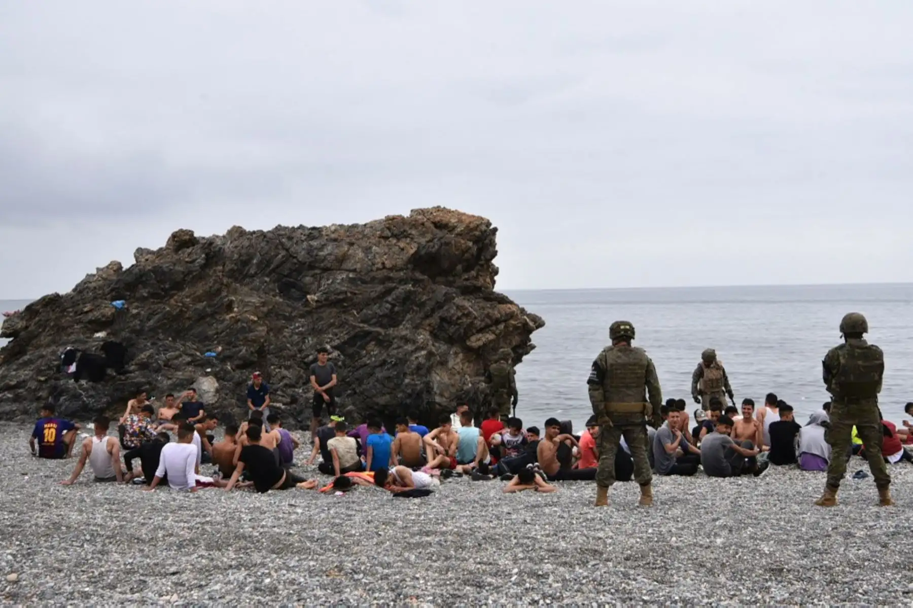 Los migrantes que llegaron nadando al enclave español de Ceuta, descansan mientras los soldados españoles hacen guardia el 18 de mayo de 2021 en Ceuta.  España ha devuelto a Marruecos casi la mitad de los 6.000 migrantes que entraron en su enclave de Ceuta, mientras cientos más intentaban entrar en su otro territorio del norte de África. Foto: AFP