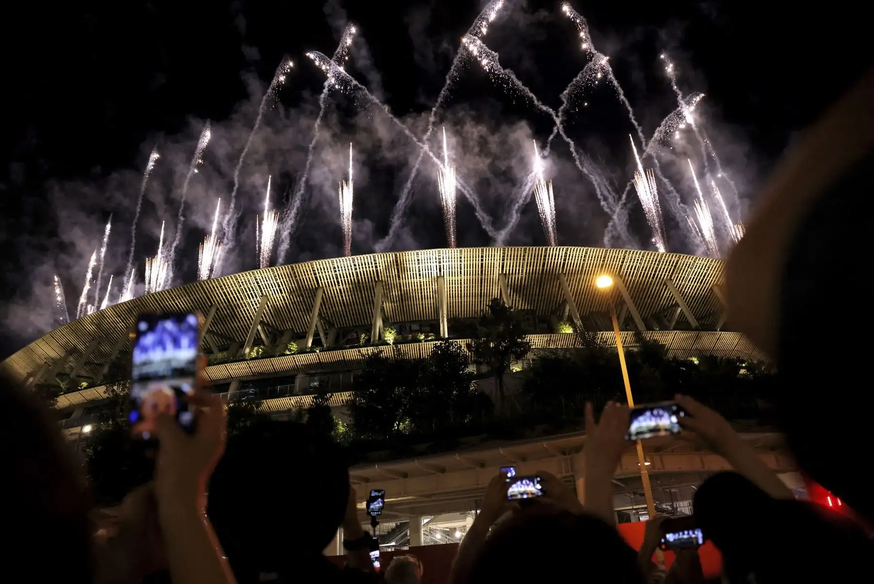 Personas afuera del Estadio Olímpico toman fotografías de los fuegos artificiales que iluminan el cielo durante la ceremonia de clausura de los Juegos Olímpicos de Tokio 2020. Foto: AFP