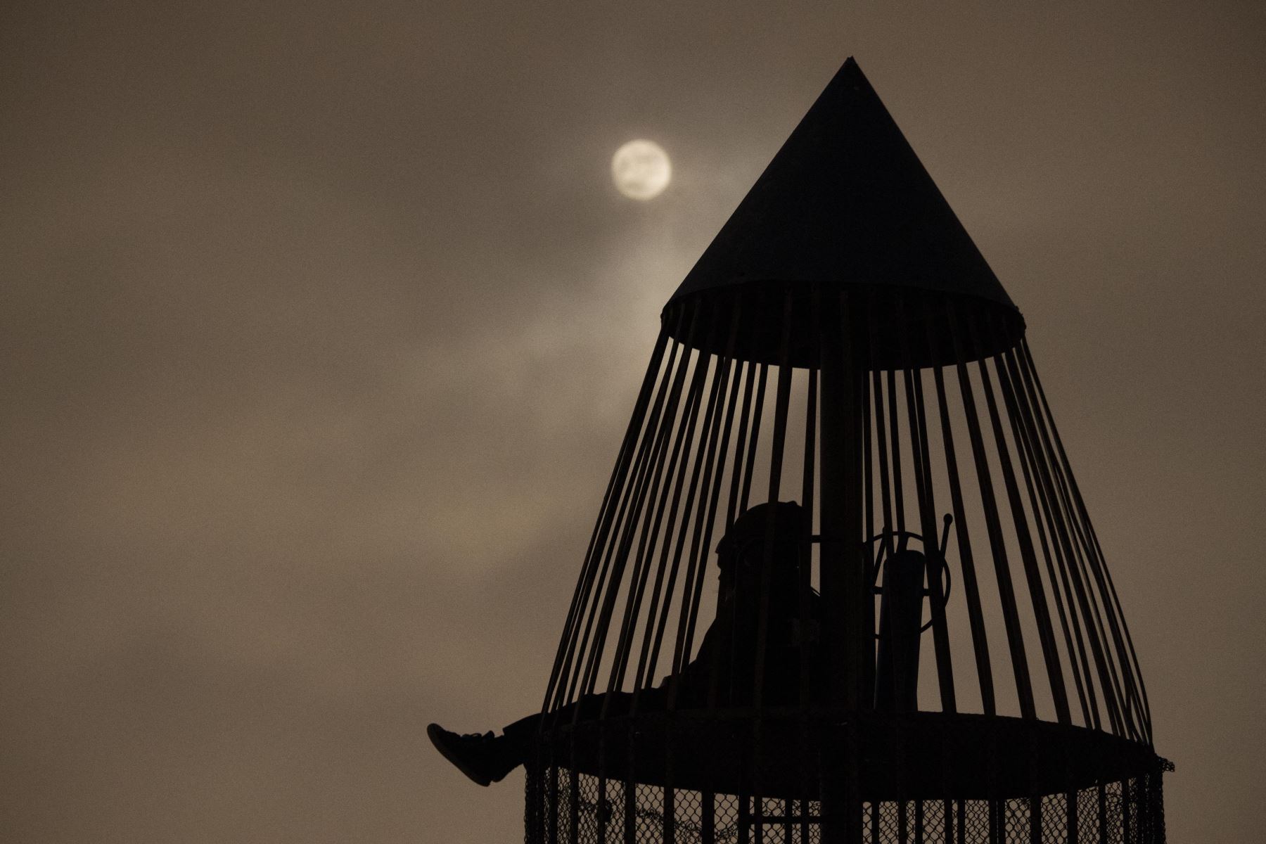 La luna brilla a través de las nubes mientras las personas se sientan dentro de una torre de juegos con temática de cohetes antes de un eclipse lunar. Foto: AFP