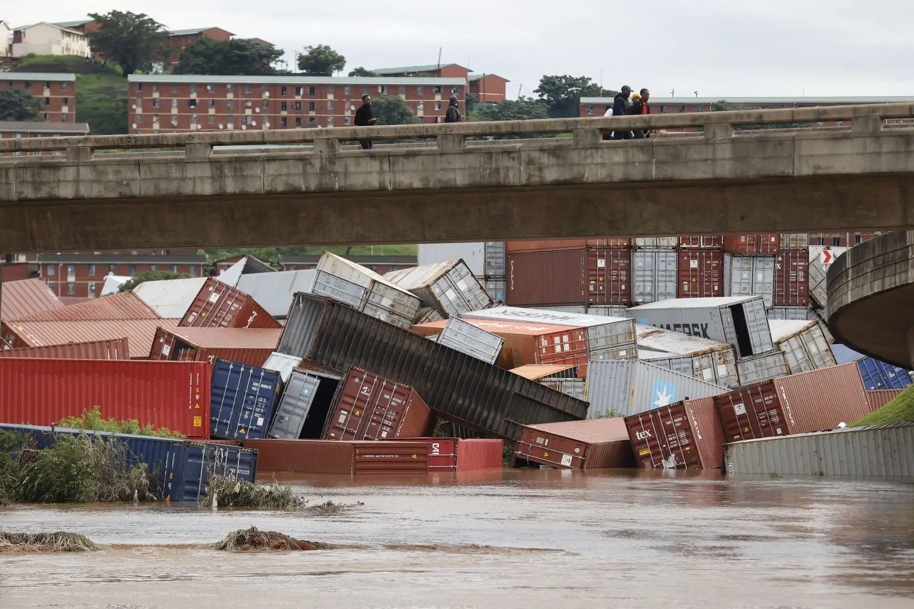 Los residentes del municipio de Umlazi se paran sobre un puente y observan los contenedores que se cayeron en una instalación de almacenamiento de contenedores luego de fuertes lluvias y vientos en Durban, el 12 de abril de 2022. 
Foto: ANDINA/AFP