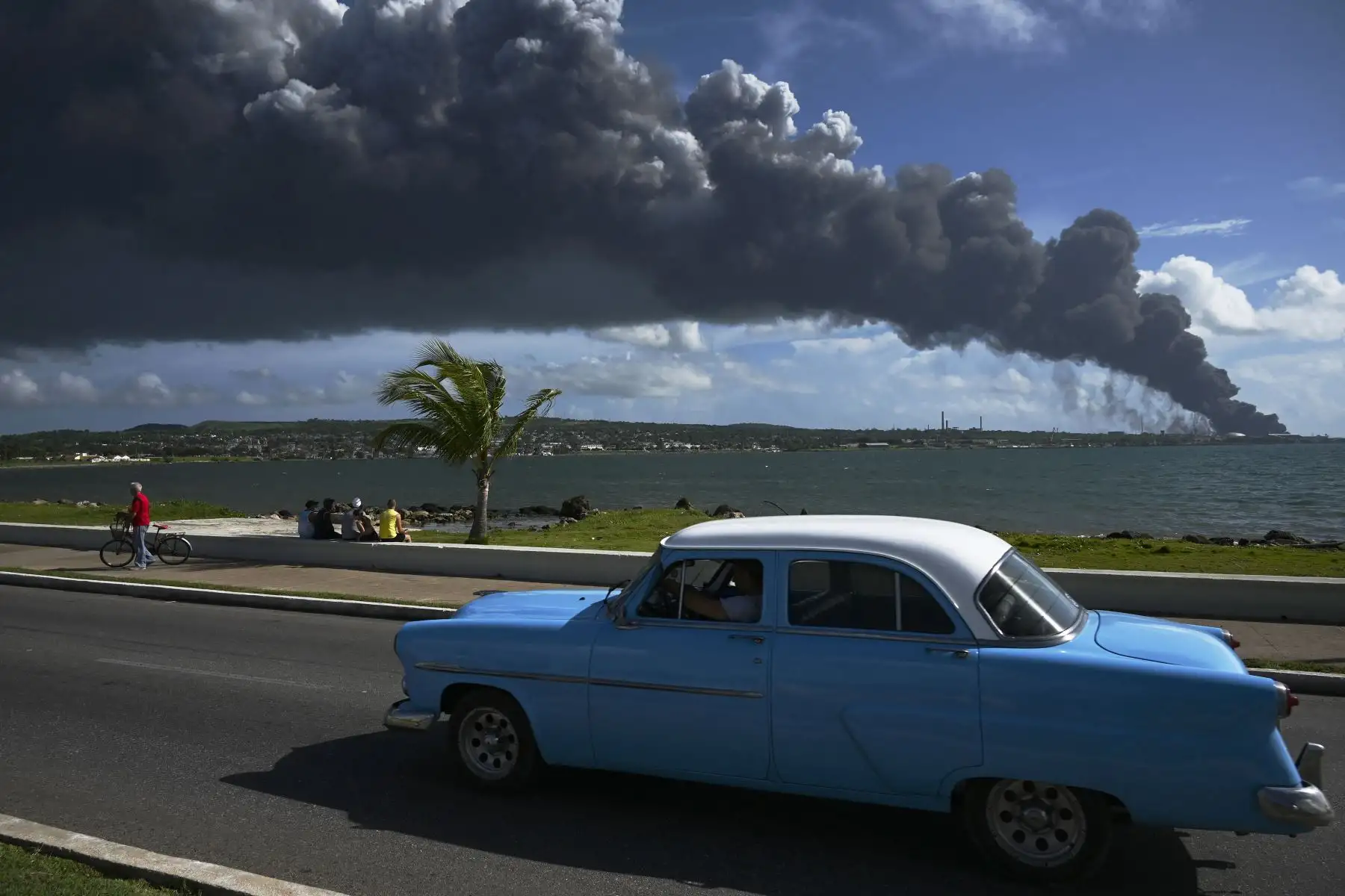 Se ve humo negro de un tanque de petróleo en llamas mientras un viejo automóvil estadounidense pasa por una calle en Matanzas, Cuba. El incendio provocado por un rayo el viernes en un depósito de combustible en Matanzas, en el oeste de Cuba, se extendió a un segundo tanque.
Foto: AFP