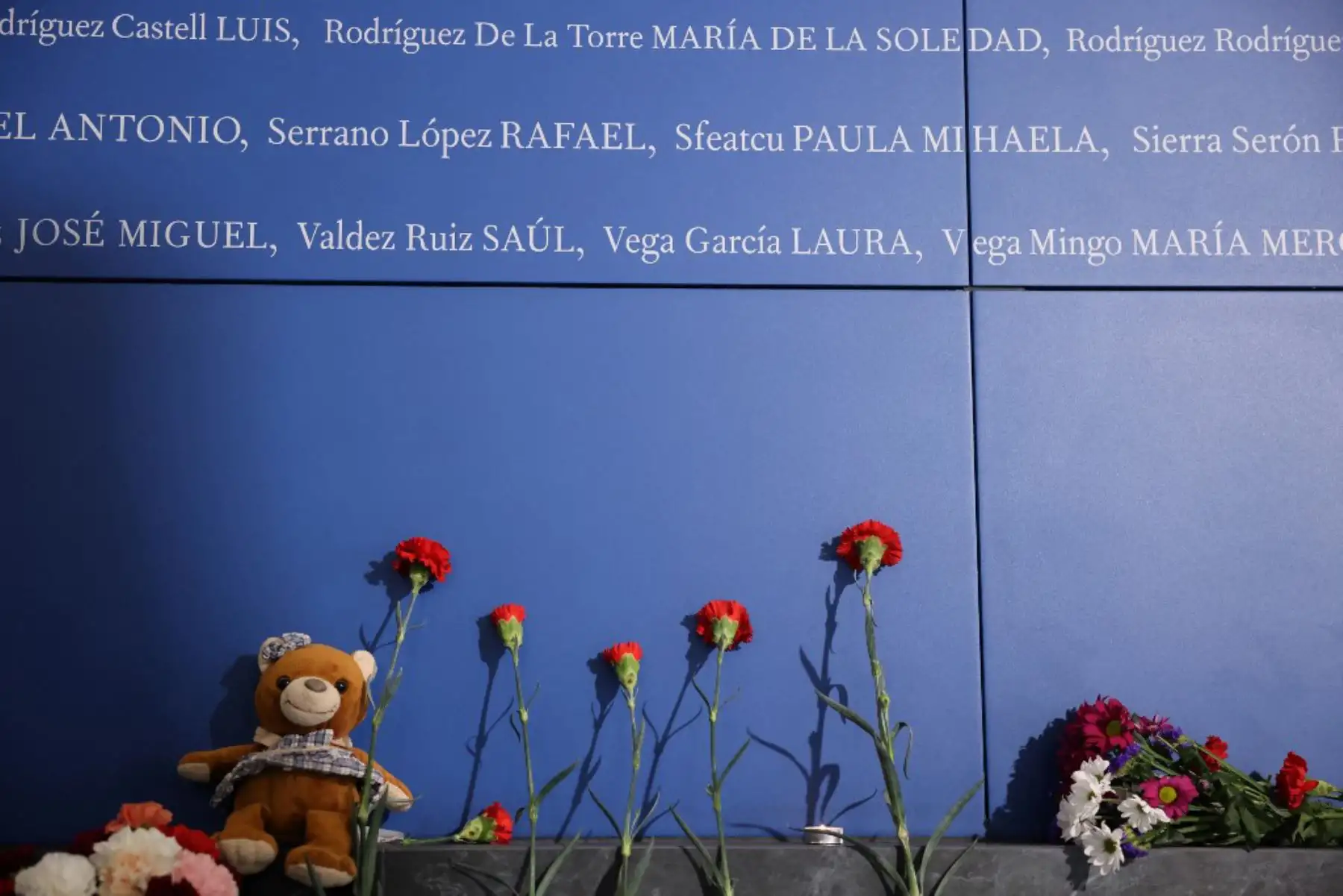 Flores de clavel y un osito de peluche colocados en la parte inferior de un panel con los nombres de las víctimas de los atentados del 11-M, que mataron a 192 personas el 11 de marzo de 2004. Foto: AFP