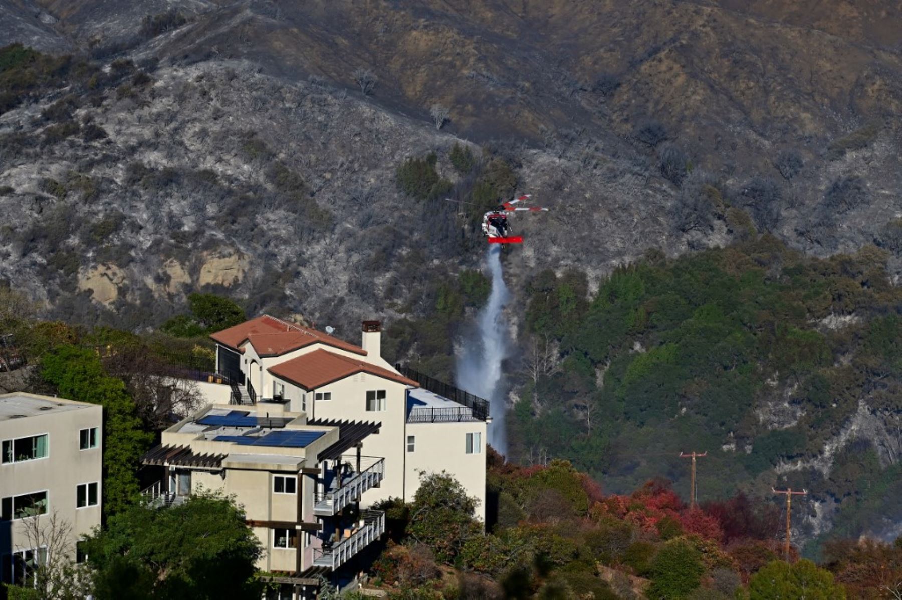 Un helicóptero CalFire deja caer agua junto a un edificio cerca de las laderas quemada por el incendio de Palisades en Fernwood, Topanga, una comunidad en el oeste del condado de Los Ángeles, California. Foto: AFP