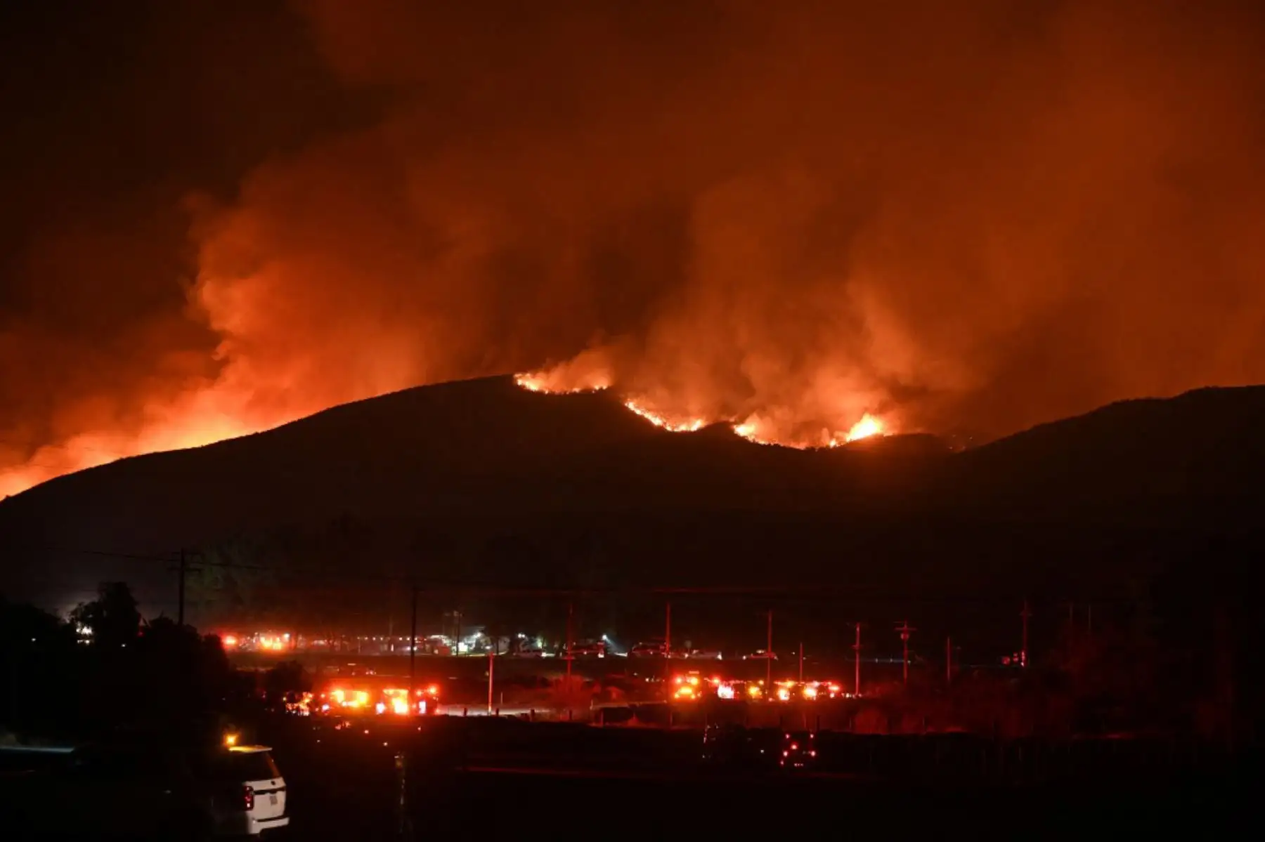 Camiones de bomberos y vehículos de seguridad pública se ven en primer plano mientras llamas rojas brillantes y humo se elevan desde el incendio Hughes en Castaic en el noroeste del condado de Los Ángeles, California. Foto: AFP