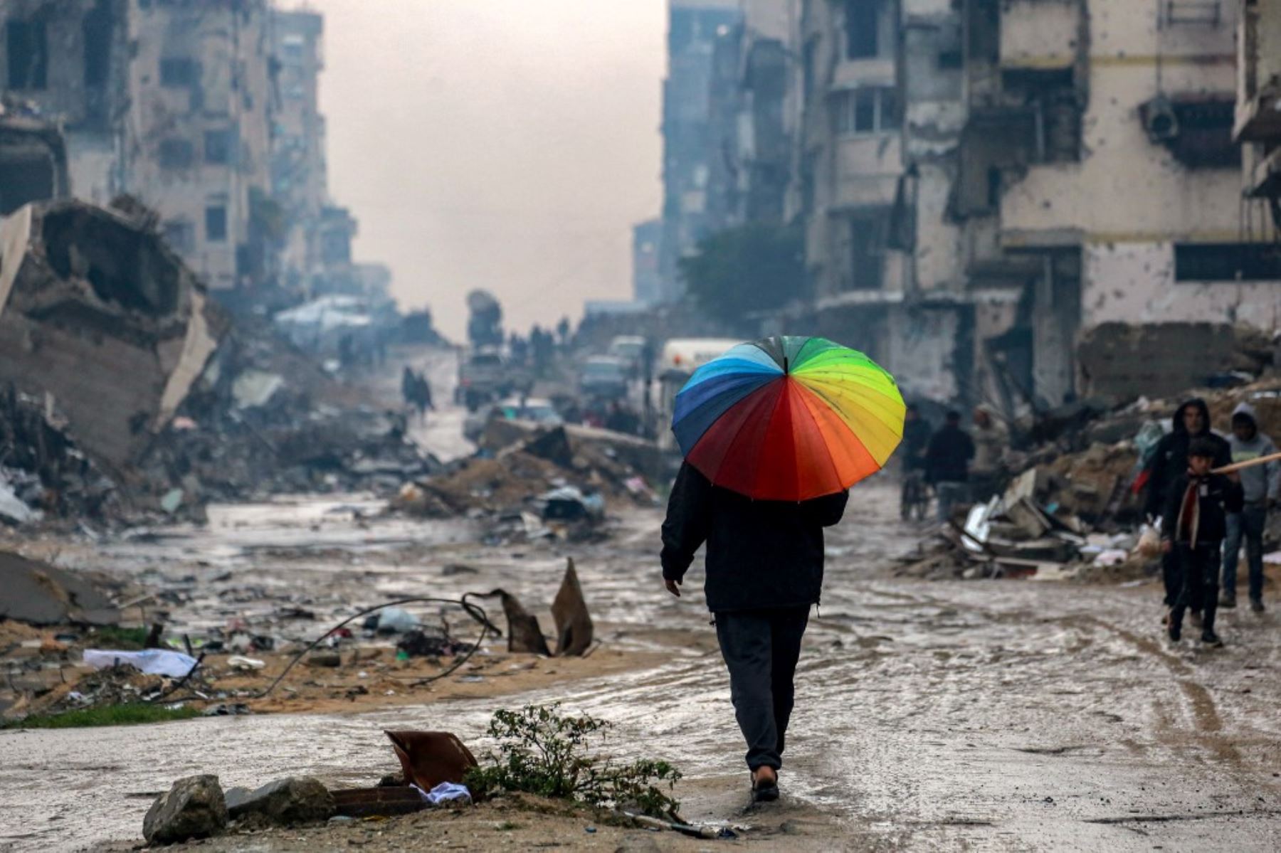 Un hombre camina con un paraguas de color arcoíris por una calle embarrada pasando los escombros de edificios destruidos y muy dañados en el norte de la ciudad de Gaza el 10 de febrero de 2025 en medio del actual acuerdo de alto el fuego en la guerra entre Israel y Hamas. (Foto de BASHAR TALEB / AFP)