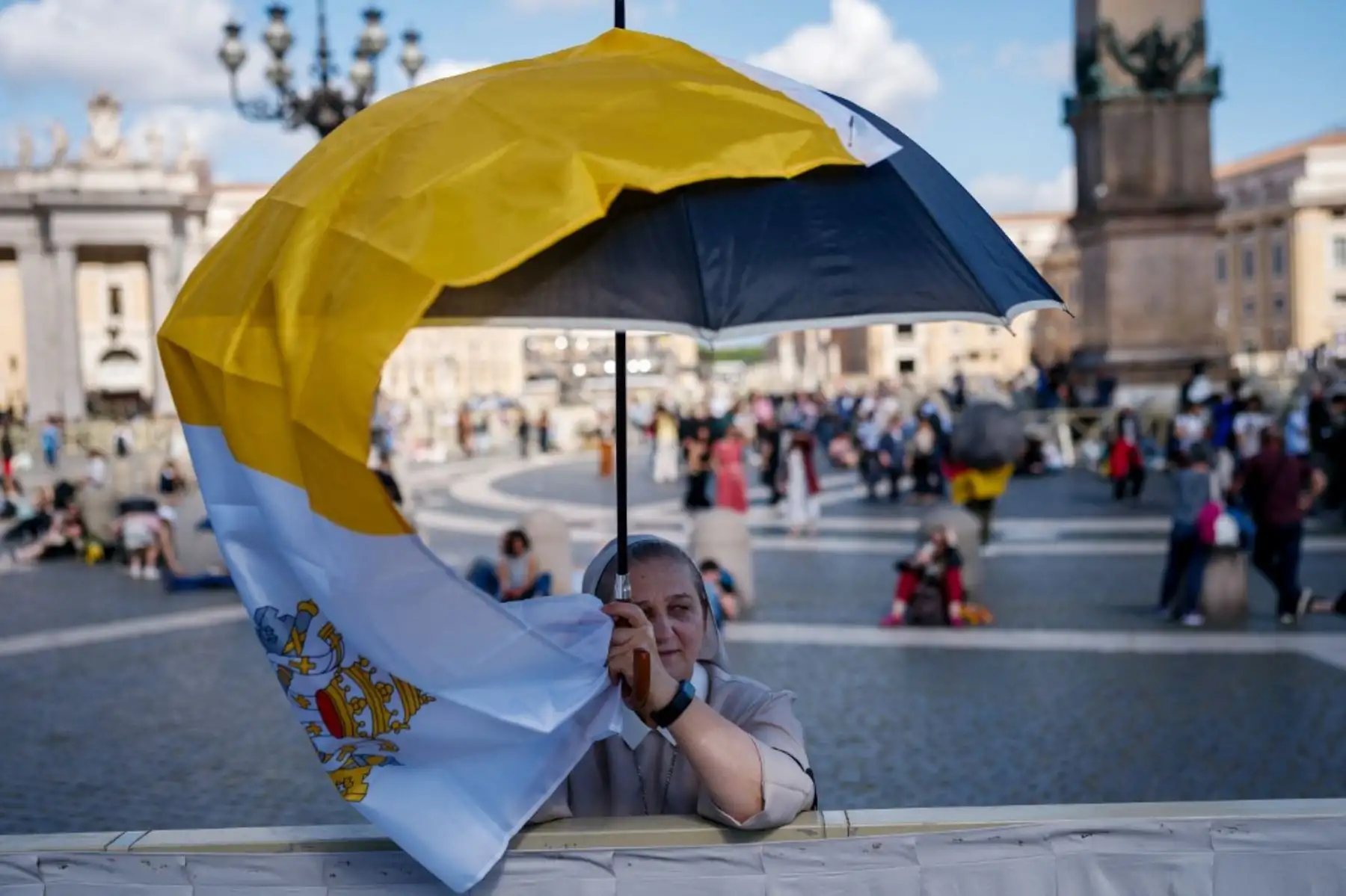 Una monja se protege del sol con una bandera del Vaticano y un paraguas mientras espera en la Plaza de San Pedro a que el humo salga de la chimenea de la Capilla Sixtina. Foto: AFP