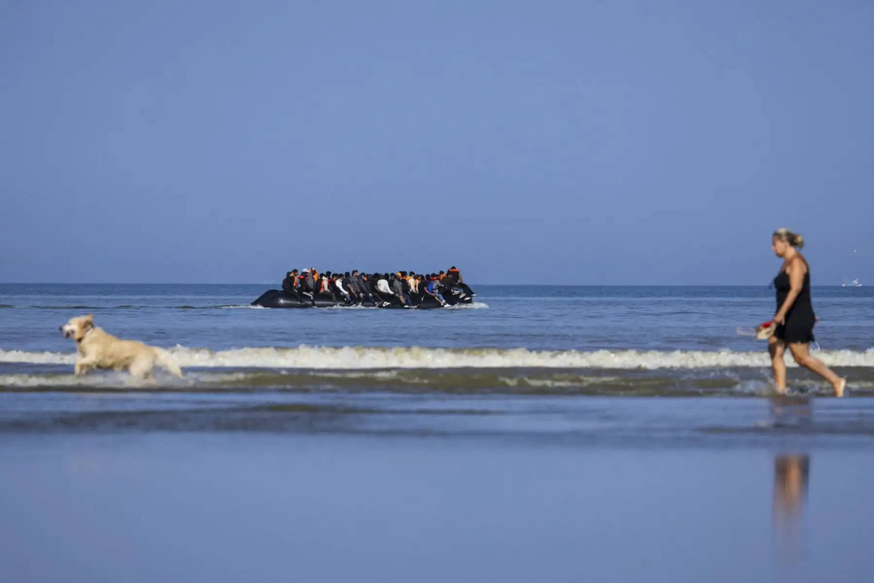 Una turista pasea a su perro mientras migrantes se acercan en el barco de un contrabandista en un intento de cruzar el Canal de la Mancha fuera de la playa de Hardelot en Neufchatel-Hardelot, norte de Francia, el 30 de junio de 2025. (Foto de Sameer Al-DOUMY / AFP)