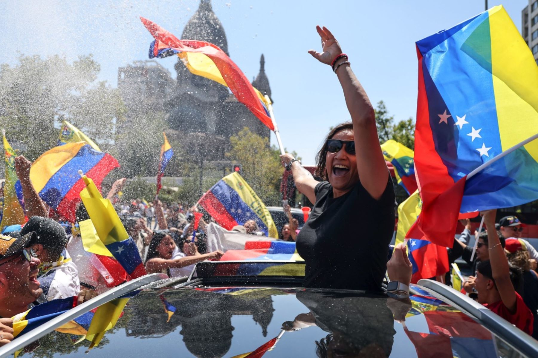 Una mujer celebra desde el techo de un vehículo mientras venezolanos agitan banderas en las calles de Santiago, tras conocerse la captura de Nicolás Maduro por fuerzas estadounidenses. La escena, marcada por bocinas, agua lanzada al aire y gestos de euforia, refleja la intensidad emocional de la diáspora venezolana en Chile. Foto; AFP