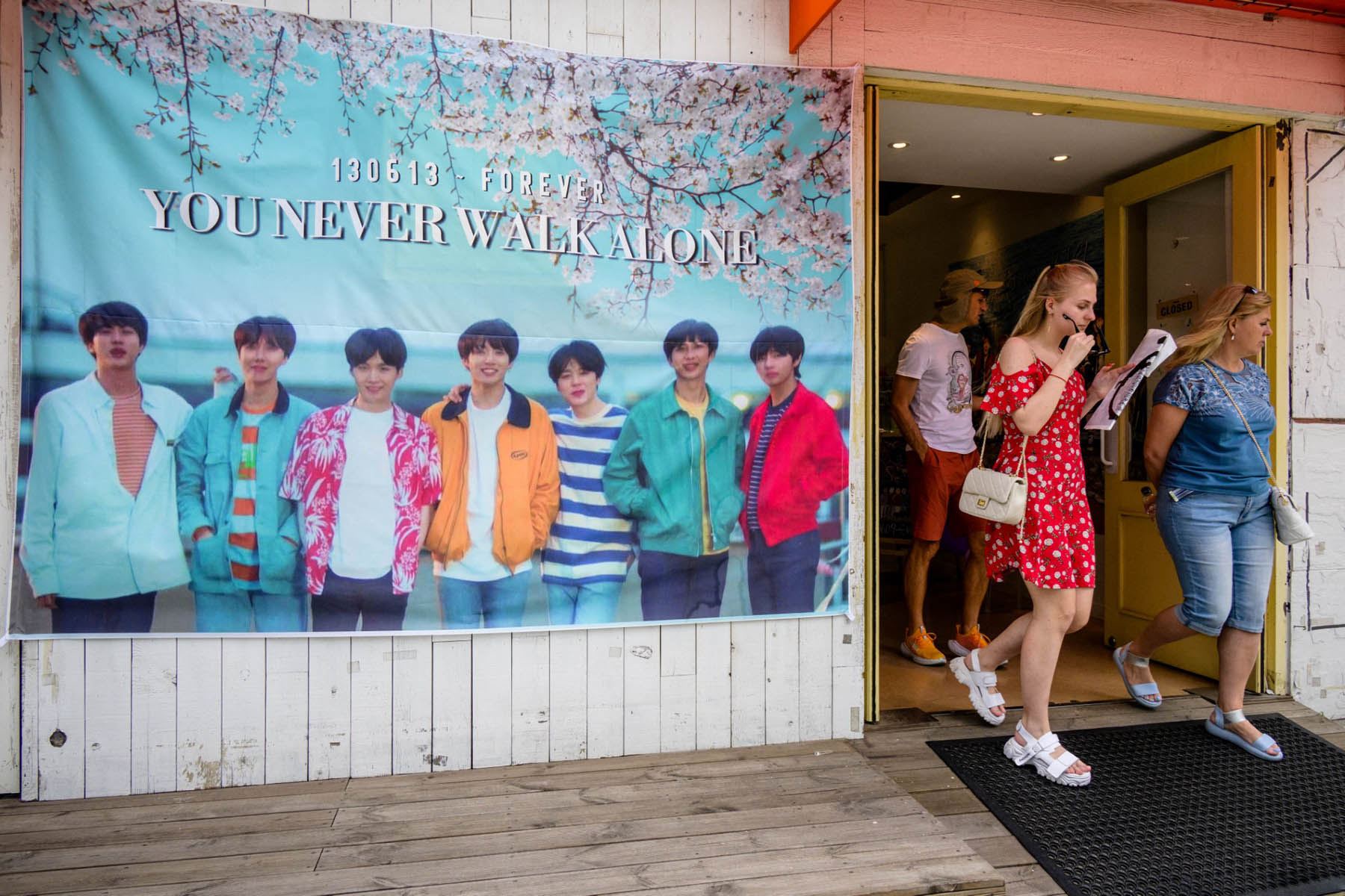 Clientes salen de una cafetería con un póster de las megaestrellas del K-pop BTS expuesto en el exterior del local en Seúl el 12 de junio de 2023, con motivo del décimo aniversario del supergrupo. Los fans de las megaestrellas del K-pop BTS acudieron en masa a los principales puntos de interés de Seúl el 12 de junio. Foto: AFP