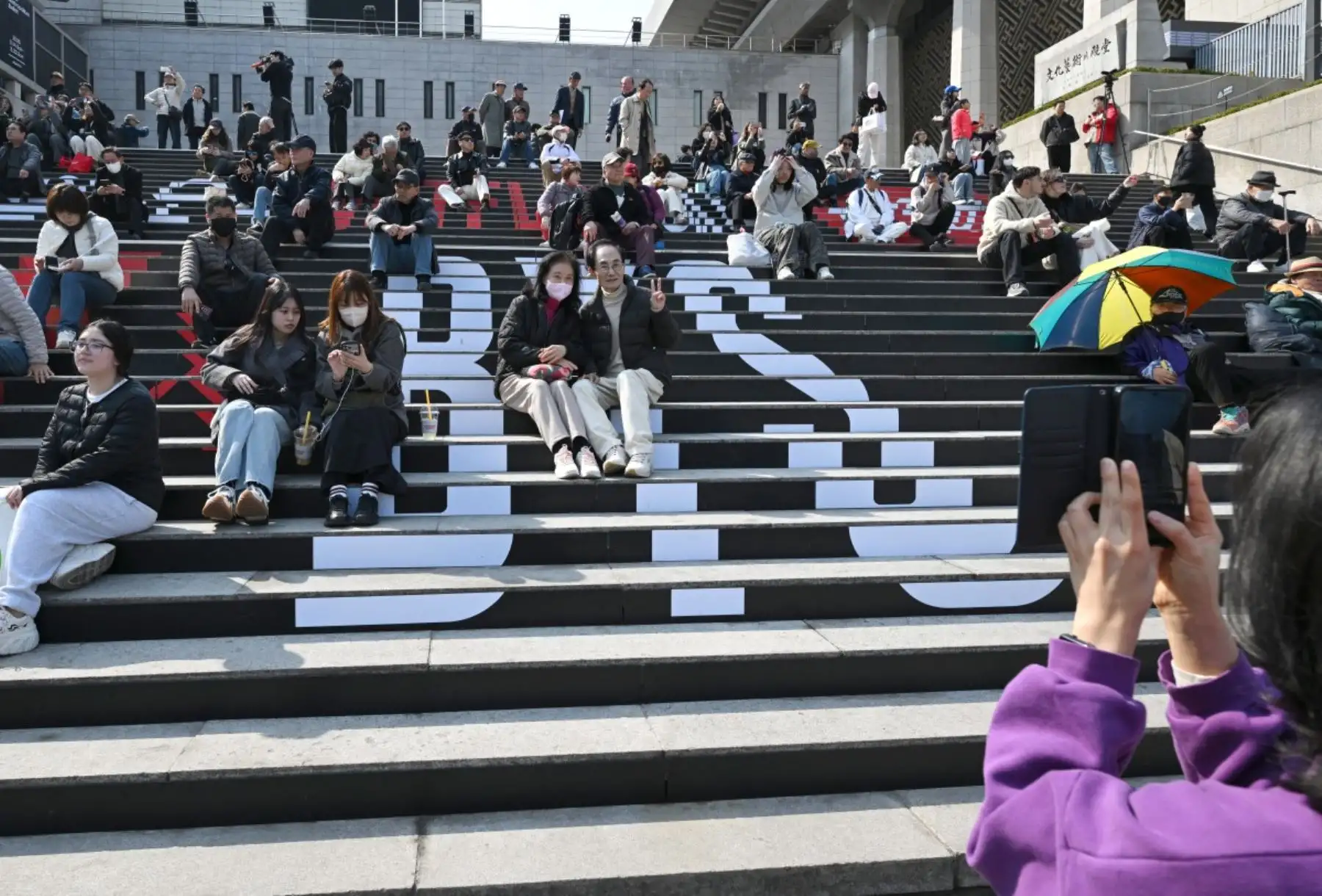 Varias personas se sientan en las escaleras que muestran el logo de BTS cerca del lugar del concierto de regreso del grupo de K-pop BTS en la plaza Gwanghwamun de Seúl. Foto: AFP
