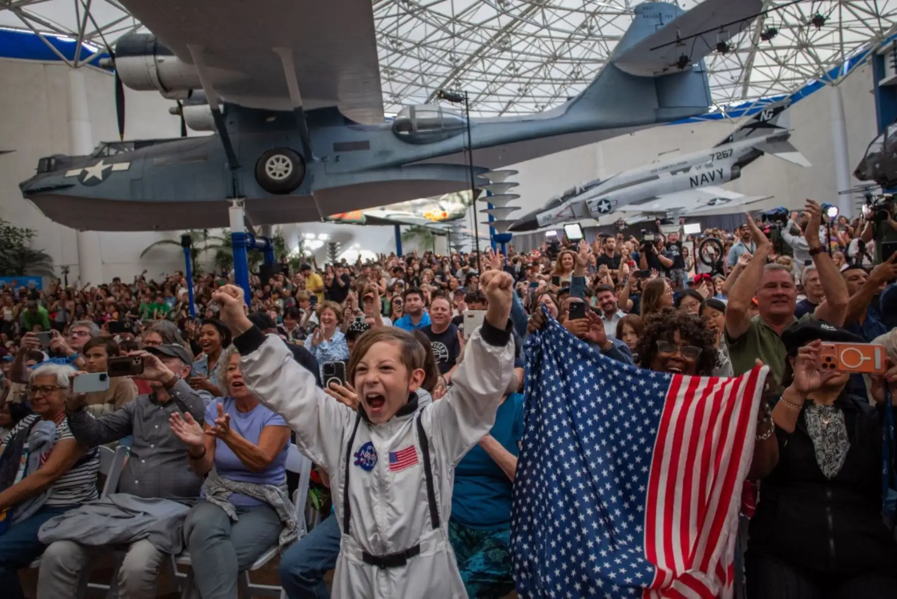 Un niño pequeño, vestido con un traje de astronauta, aparece junto a una mujer que ondea una bandera mientras observan la transmisión en vivo del regreso de la tripulación de Artemis II a la Tierra en el Museo del Aire y el Espacio de San Diego, durante una fiesta para ver el amerizaje de la tripulación en el Océano Pacífico, en San Diego, California. Foto: AFP