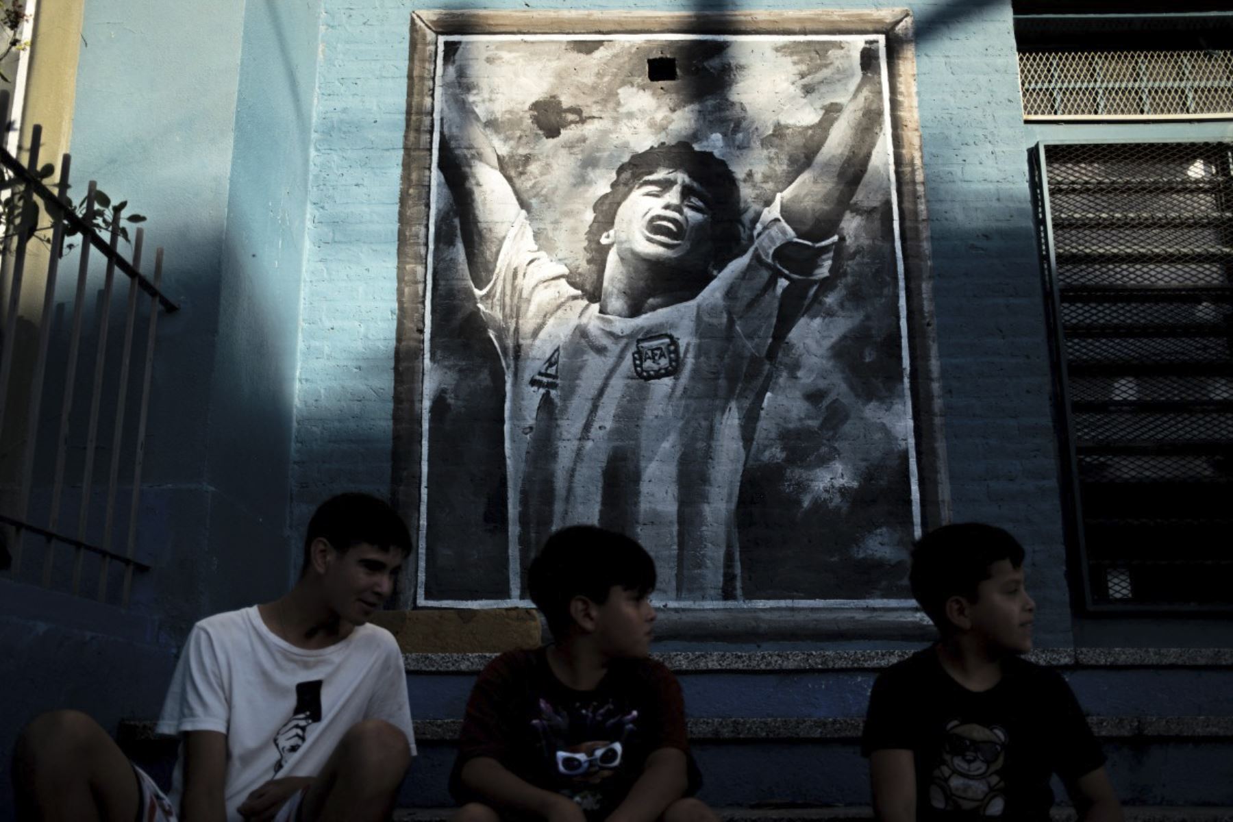 Chicos se sientan frente a un mural que representa a la difunta estrella del fútbol Diego Maradona en el barrio de su ciudad natal, Villa Fiorito, en la provincia de Buenos Aires, Argentina, el 9 de abril de 2026. Foto: AFP