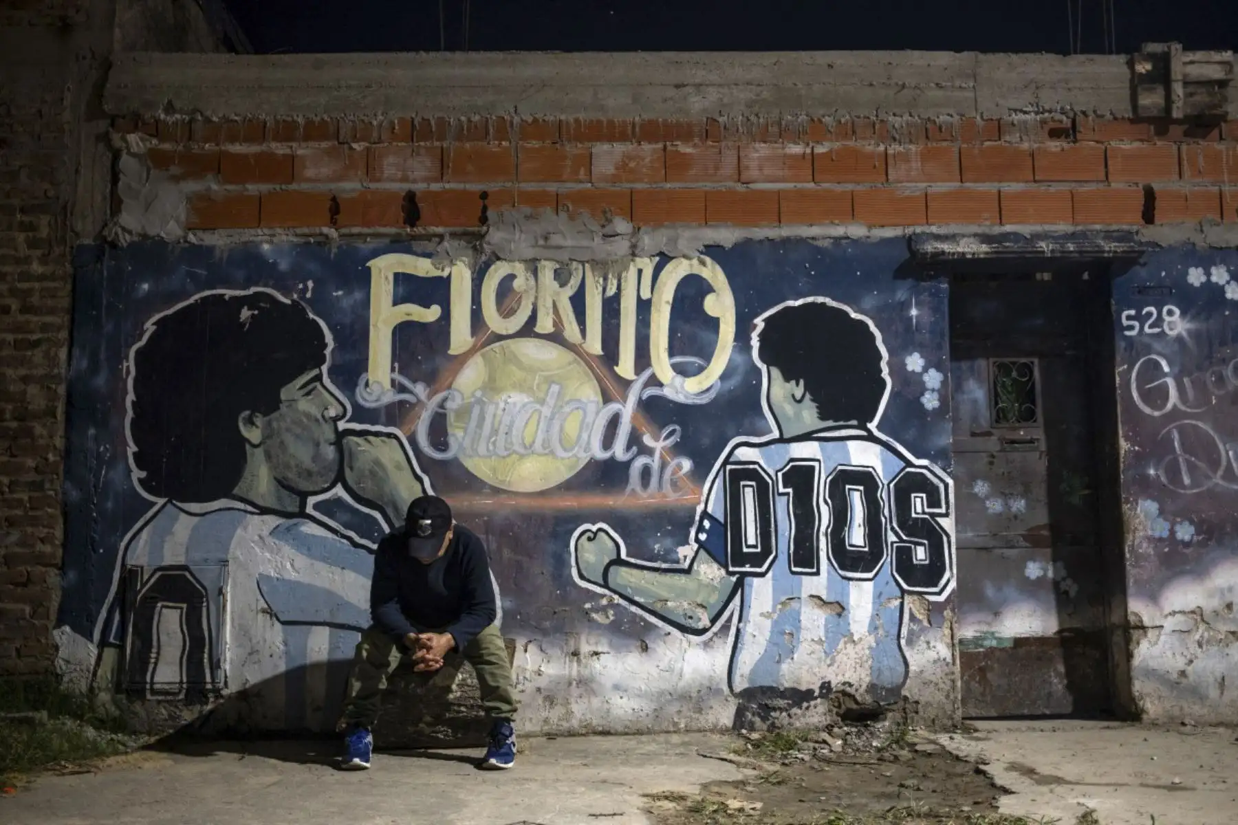 Un hombre se sienta frente a un mural que representa a la difunta estrella del fútbol Diego Maradona y leyendo en español "Ciudad de Dios de Fiorito" en el patio de la casa de la infancia de la difunta estrella del fútbol Diego Maradona, ahora transformada en un comedor de beneficencia para vecinos necesitados, en Villa Fiorito, provincia de Buenos Aires, Argentina, el 9 de abril de 2026. Foto: AFP