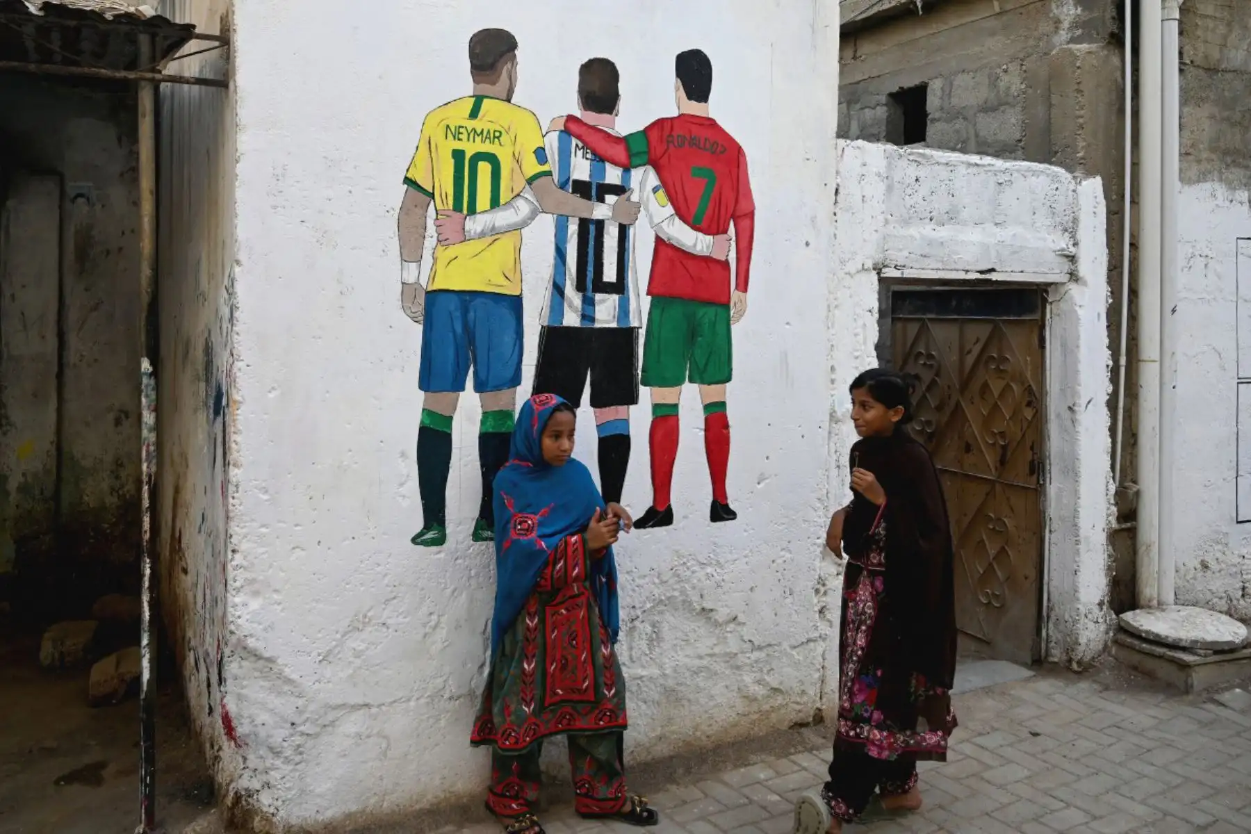 Unas niñas pasan junto a un mural con las imágenes de los futbolistas Neymar, Lionel Messi  y Cristiano Ronaldo antes de la Copa Mundial de la FIFA 2026. El mural se exhibe en una calle de Karachi, Pakistán. Foto: AFP