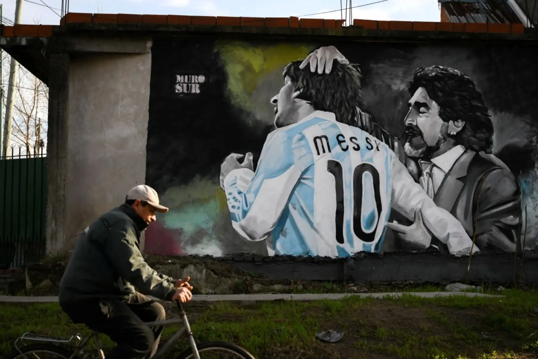 Un hombre pasa en bicicleta junto a un mural que representa al fallecido legendario futbolista argentino Diego Maradona y al delantero estrella Lionel Messi durante un partido del Mundial de 2010, en Villa Fiorito, en las afueras de Buenos Aires, el 14 de julio de 2026. Foto: AFP