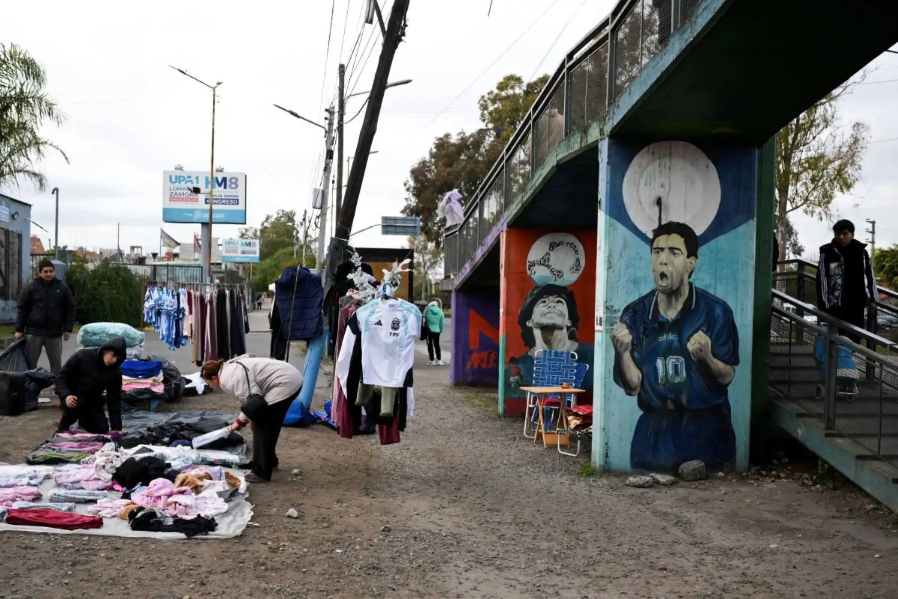 Vendedores ofrecen ropa a la venta cerca de murales que representan al fallecido legendario futbolista argentino Diego Maradona en Villa Fiorito, en las afueras de Buenos Aires, el 14 de julio de 2026. Foto: AFP