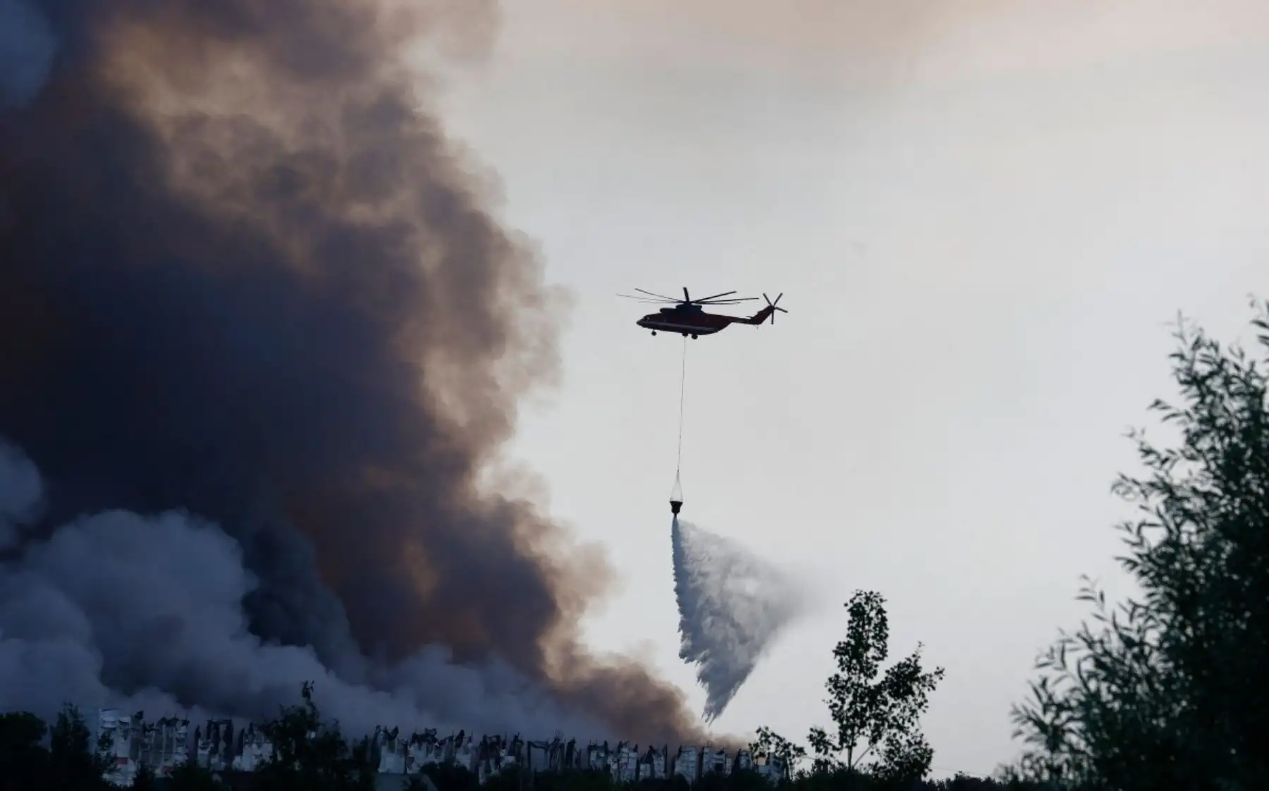 Un helicóptero de extinción de incendios intenta sofocar un fuego en los complejos logísticos de la empresa rusa de comercio electrónico Wildberries, en la localidad de Elektrostal (a las afueras de Moscú), el 18 de julio de 2026.
Un ataque con drones ucranianos contra dos centros logísticos en Rusia causó la muerte de ocho personas y dejó 24 heridas, según informaron las autoridades el 18 de julio de 2026, jornada en la que la región de Moscú reportó una nueva oleada de ataques con drones. La empresa rusa de comercio electrónico Wildberries comunicó que sus complejos logísticos en Kotovsk y Elektrostal —situados en las regiones de Tambov y Moscú, respectivamente— fueron objeto de ataques. Una persona falleció y 37 resultaron heridas en el almacén de Elektrostal, según declaró el gobernador de la región, Andréi Vorobiov. Foto: AFP