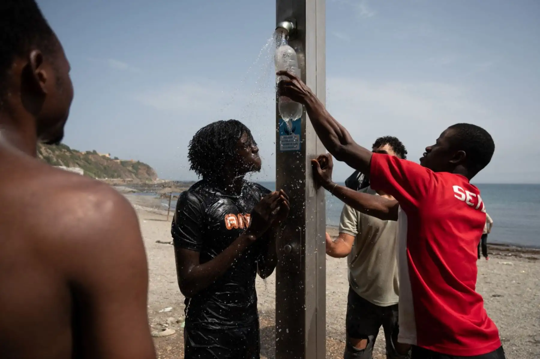 Los migrantes se refrescan bajo la ducha en la playa de Trampolín, mientras esperan para entrar al centro de acogida temporal CETI en el enclave español de Ceuta, en el norte de África, el 1 de agosto de 2026. Foto: AFP