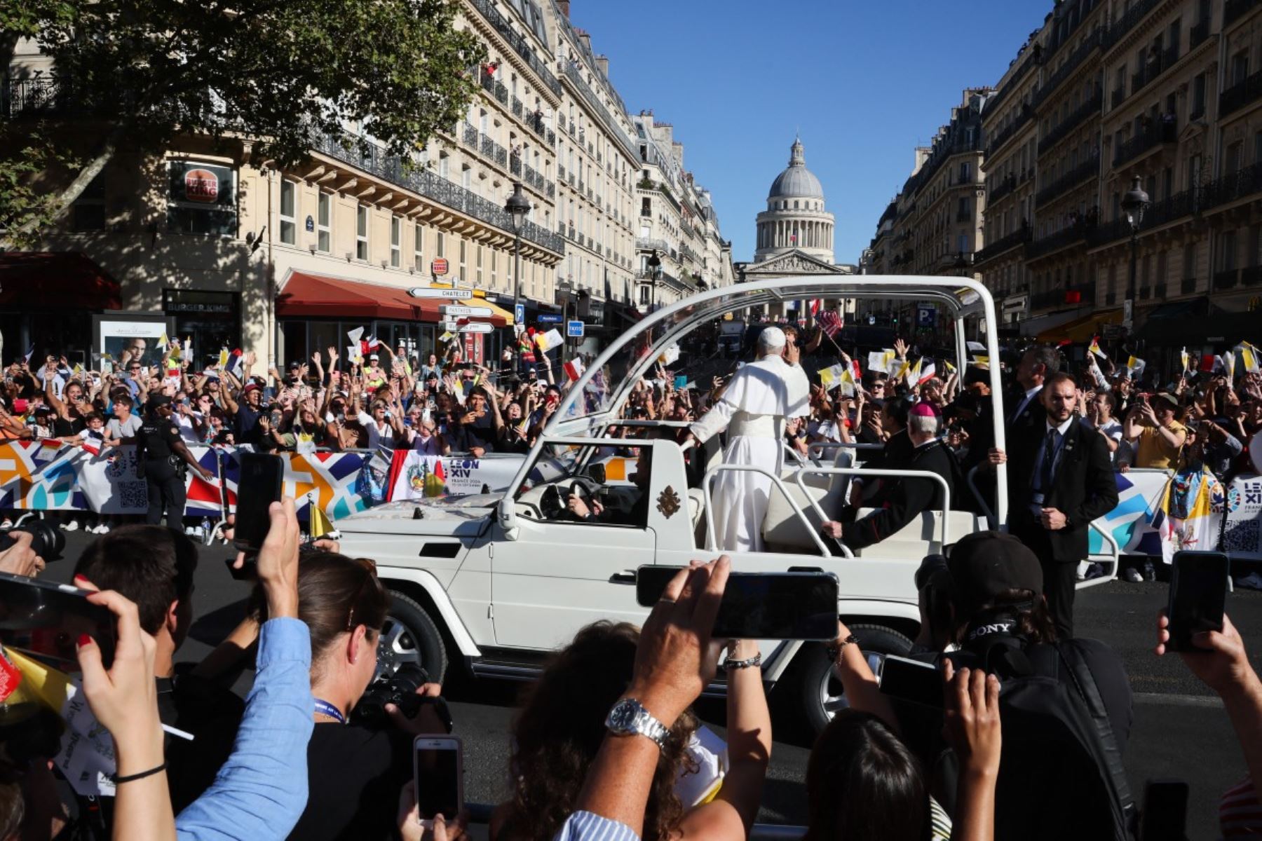 El papa León XIV, durante un recorrido por las calles de París, de pie en un papamóvil blanco descubierto y saludando a la multitud. Se observan miembros del clero, personal de seguridad, banderas y espectadores a lo largo de la calle parisina durante el recorrido papal. Foto: AFP