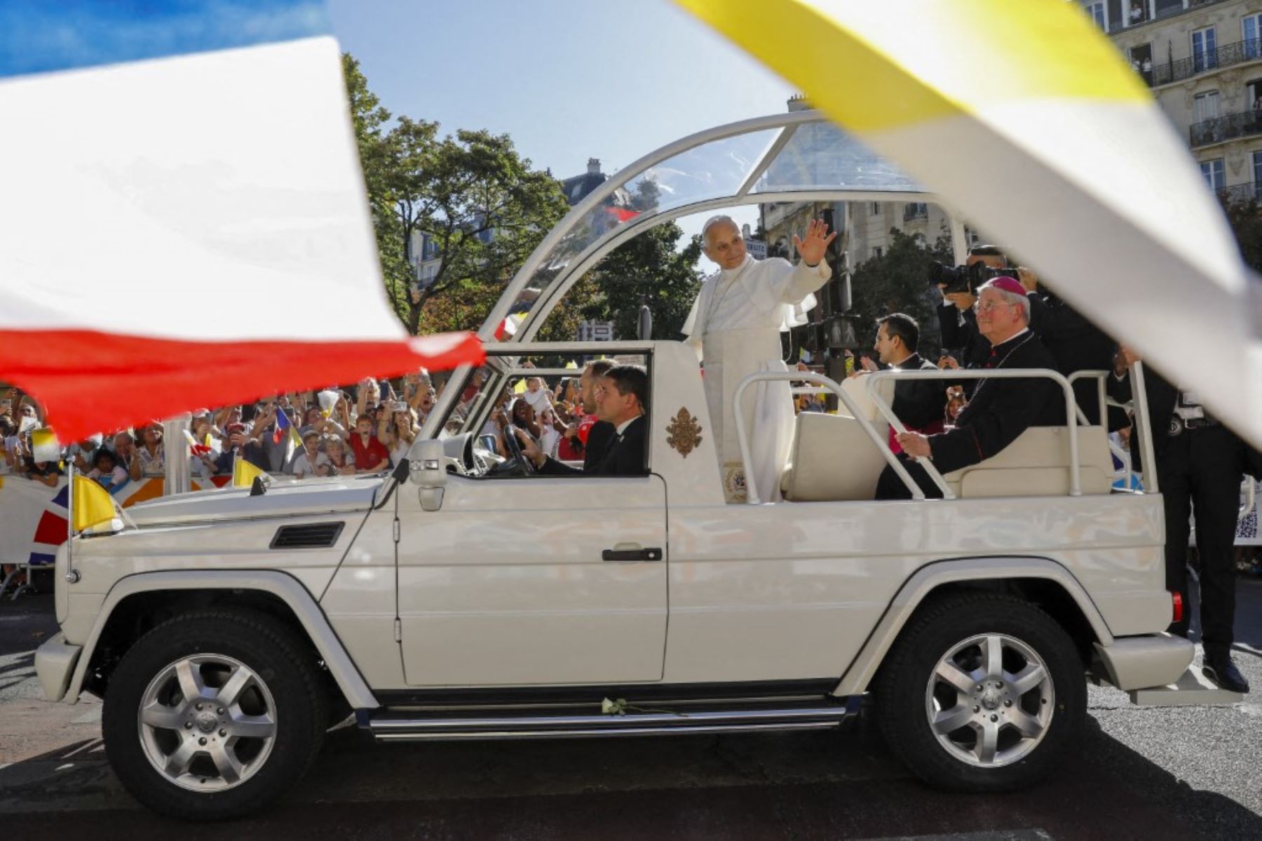 El papa León XIV, durante un recorrido por las calles de París, de pie en un papamóvil blanco descubierto y saludando a la multitud. Se observan miembros del clero, personal de seguridad, banderas y espectadores a lo largo de la calle parisina durante el recorrido papal. Foto: AFP